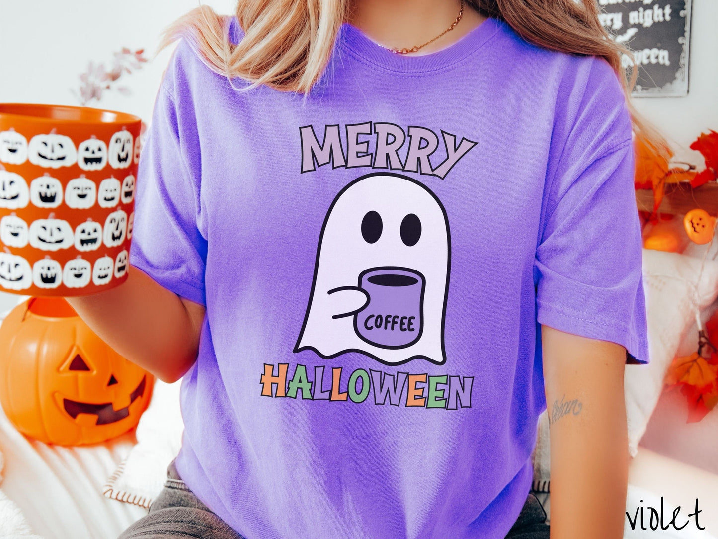 A woman wearing a cute, vintage violet colored Comfort Colors t-shirt with the text Merry Halloween in purple, orange, and green font. There is a cute ghost holding a coffee mug in between the text.