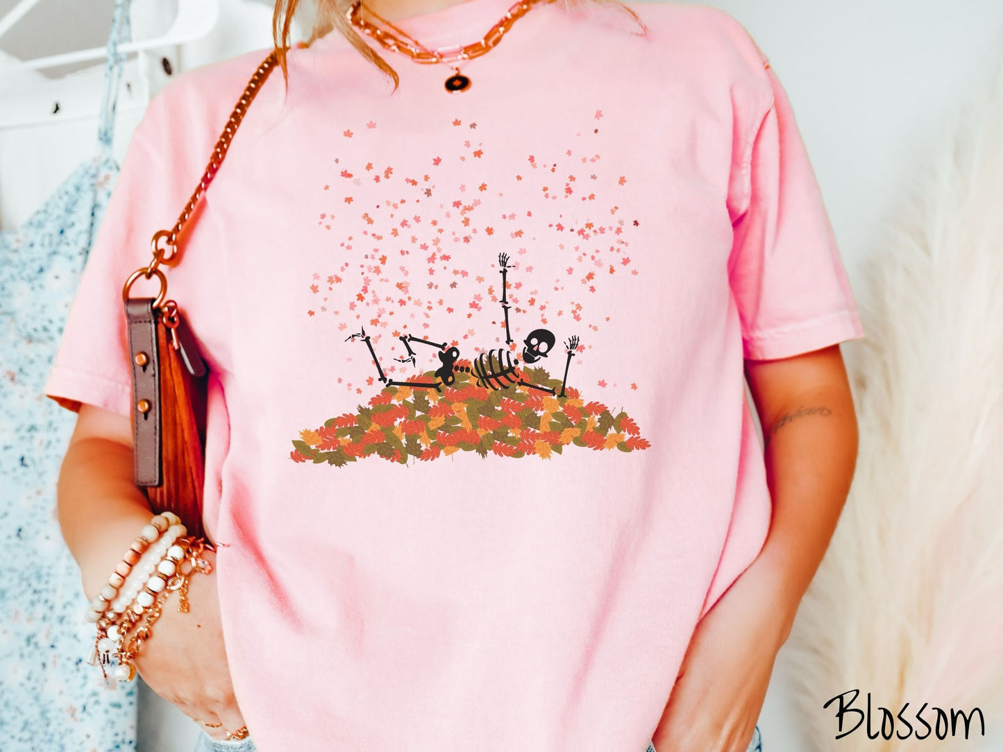 A woman wearing a cute, vintage blossom colored Comfort Colors T-shirt with a black skeleton playing in a pile of colorful fall leaves. The skeleton is throwing leaves in the air, and there are colorful leaves floating above.
