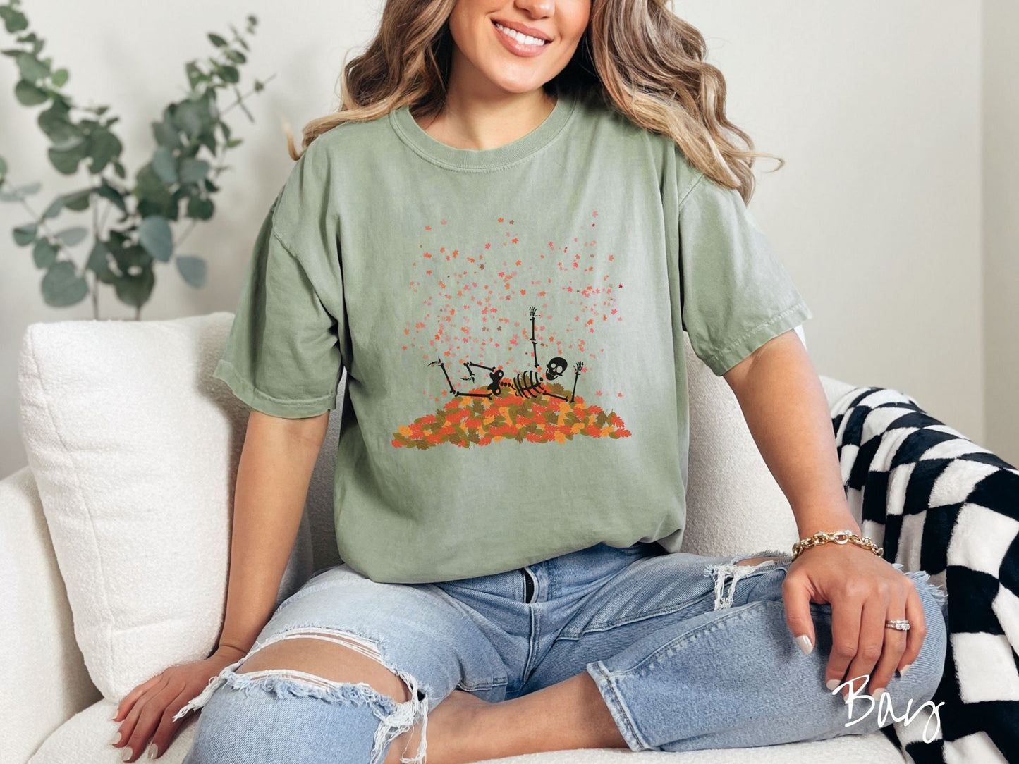 A woman wearing a cute, vintage bay colored Comfort Colors T-shirt with a black skeleton playing in a pile of colorful fall leaves. The skeleton is throwing leaves in the air, and there are colorful leaves floating above.