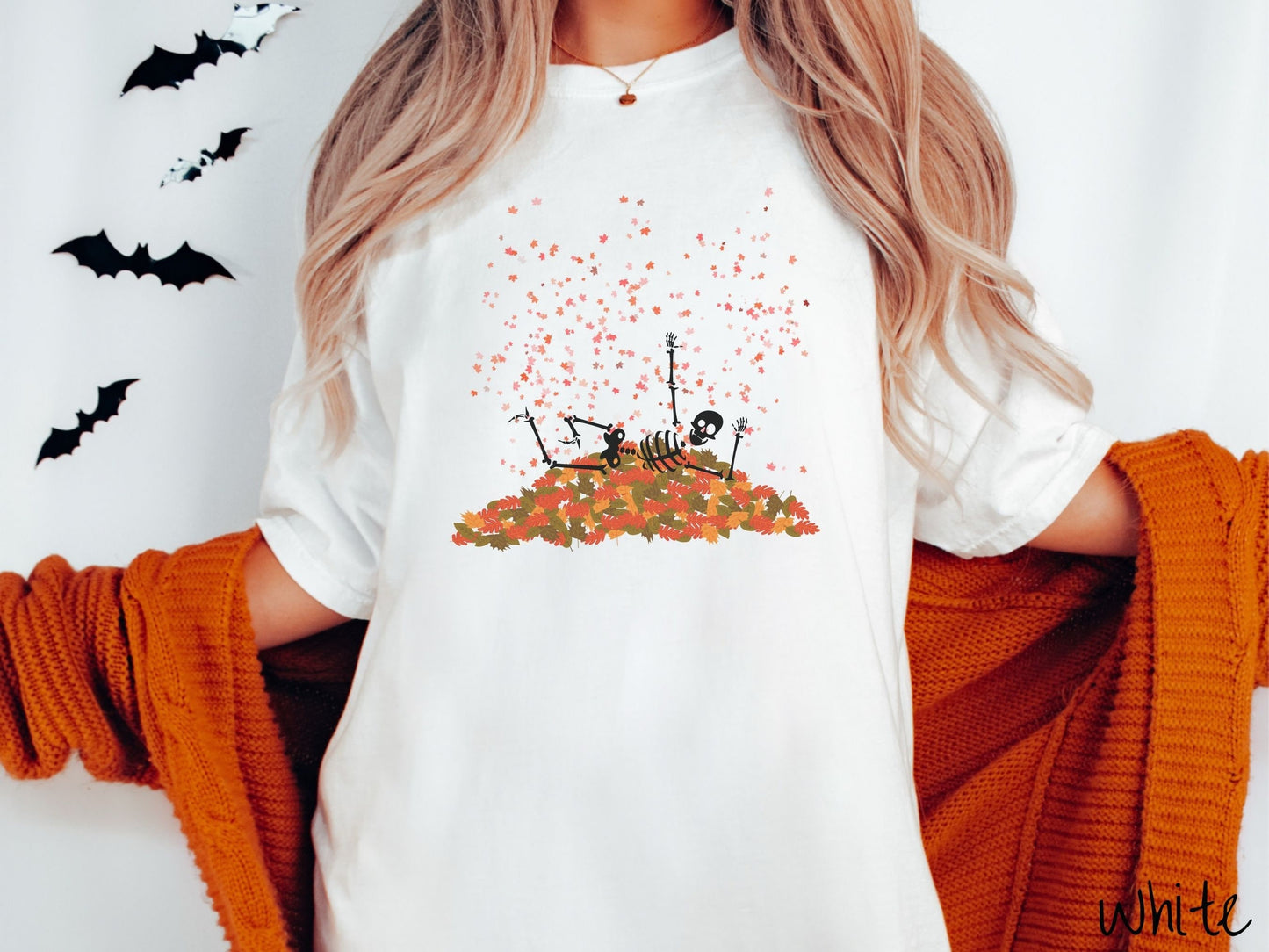A woman wearing a cute, vintage white colored Comfort Colors T-shirt with a black skeleton playing in a pile of colorful fall leaves. The skeleton is throwing leaves in the air, and there are colorful leaves floating above.