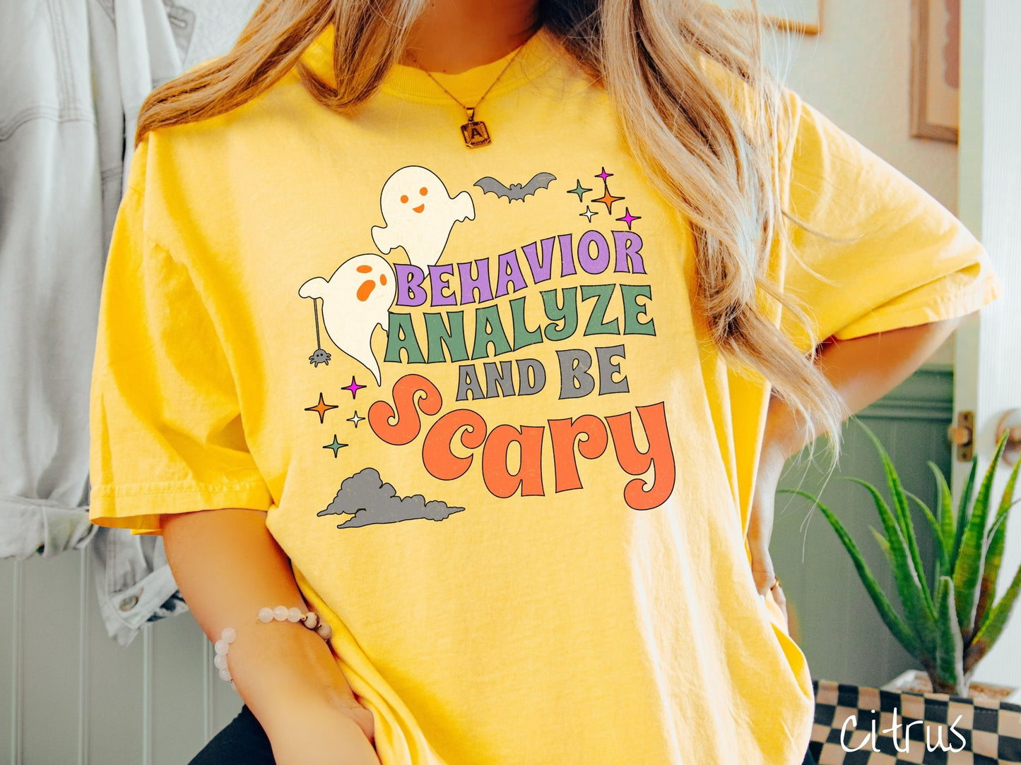 A woman wearing a cute, vintage citrus colored Comfort Colors T-shirt with the text Behavior Analyze and Be Scary in colorful font. There are orange-eyed ghosts flying above and a black bat. A black spider is dangling by its web from one ghost.