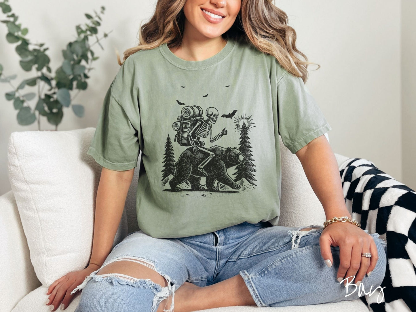 A woman wearing a cute, vintage bay colored Comfort Colors T-shirt with a smiling skeleton wearing a camping backpack and riding a bear in the woods. There are trees in the background in front of the moon and black bats in the air.