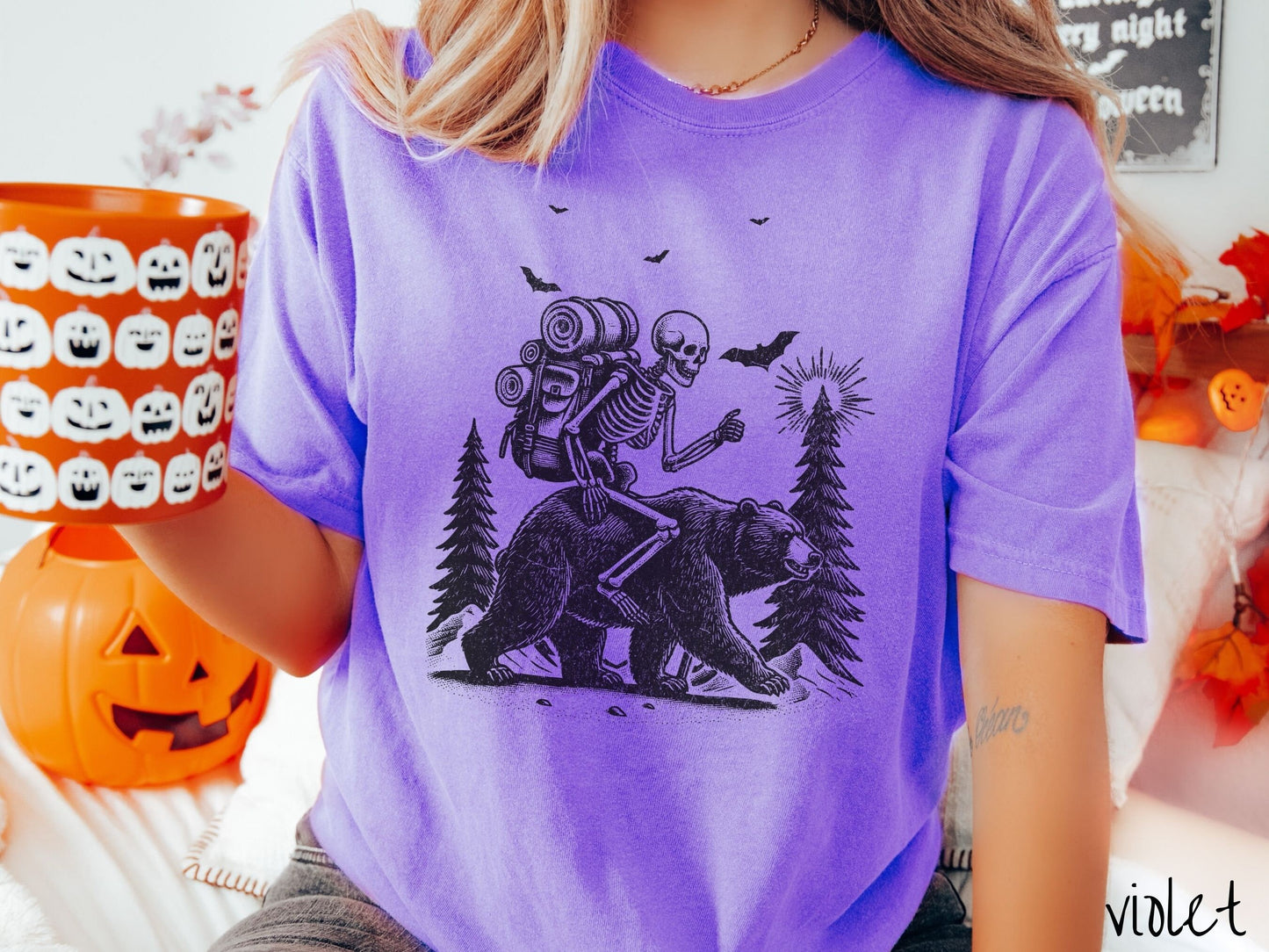 A woman wearing a cute, vintage violet colored Comfort Colors T-shirt with a smiling skeleton wearing a camping backpack and riding a bear in the woods. There are trees in the background in front of the moon and black bats in the air.