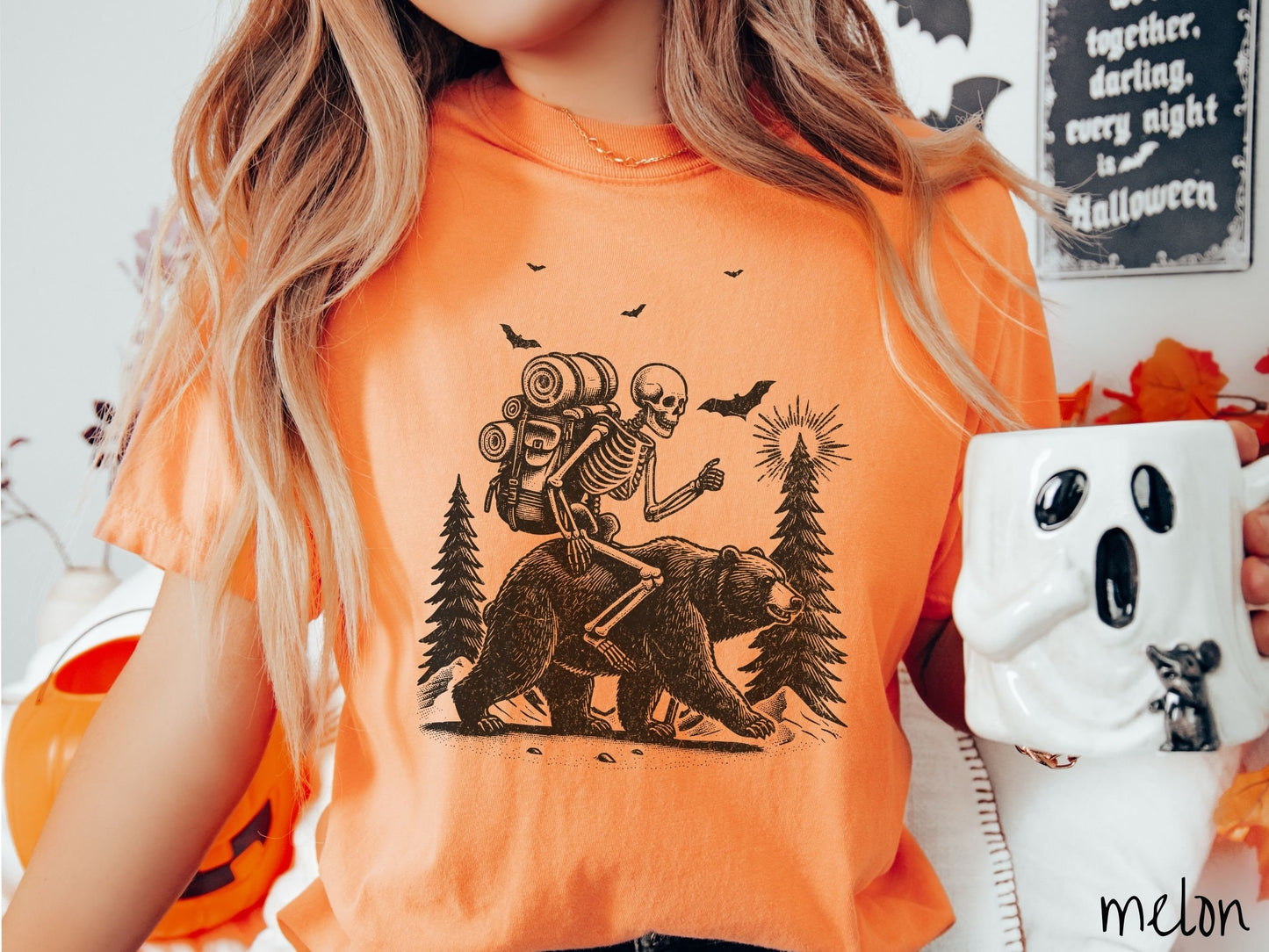 A woman wearing a cute, vintage melon colored Comfort Colors T-shirt with a smiling skeleton wearing a camping backpack and riding a bear in the woods. There are trees in the background in front of the moon and black bats in the air.