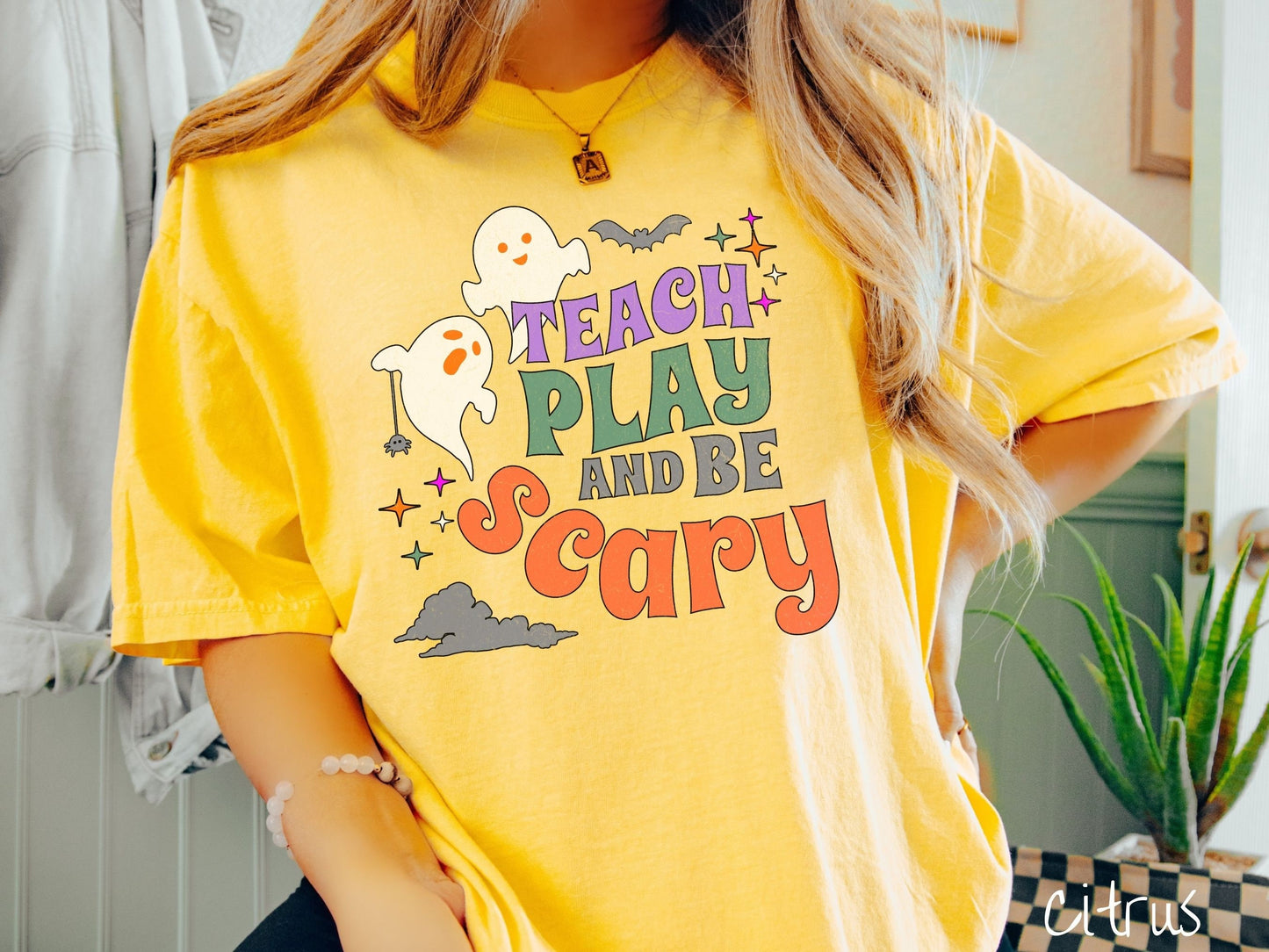 A woman wearing a cute, vintage citrus colored Comfort Colors T-shirt with the text Teach Play and Be Scary in colorful font. There are orange-eyed white ghosts flying around and black bats and twinkling stars in the sky.