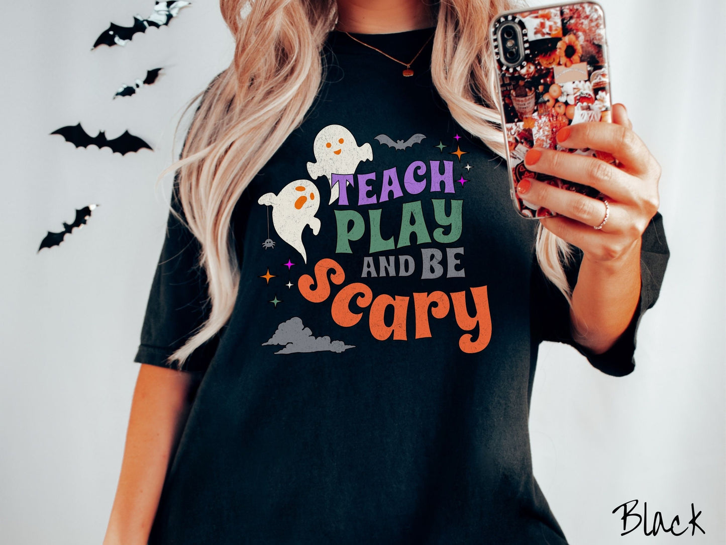 A woman wearing a cute, vintage black colored Comfort Colors T-shirt with the text Teach Play and Be Scary in colorful font. There are orange-eyed white ghosts flying around and black bats and twinkling stars in the sky.