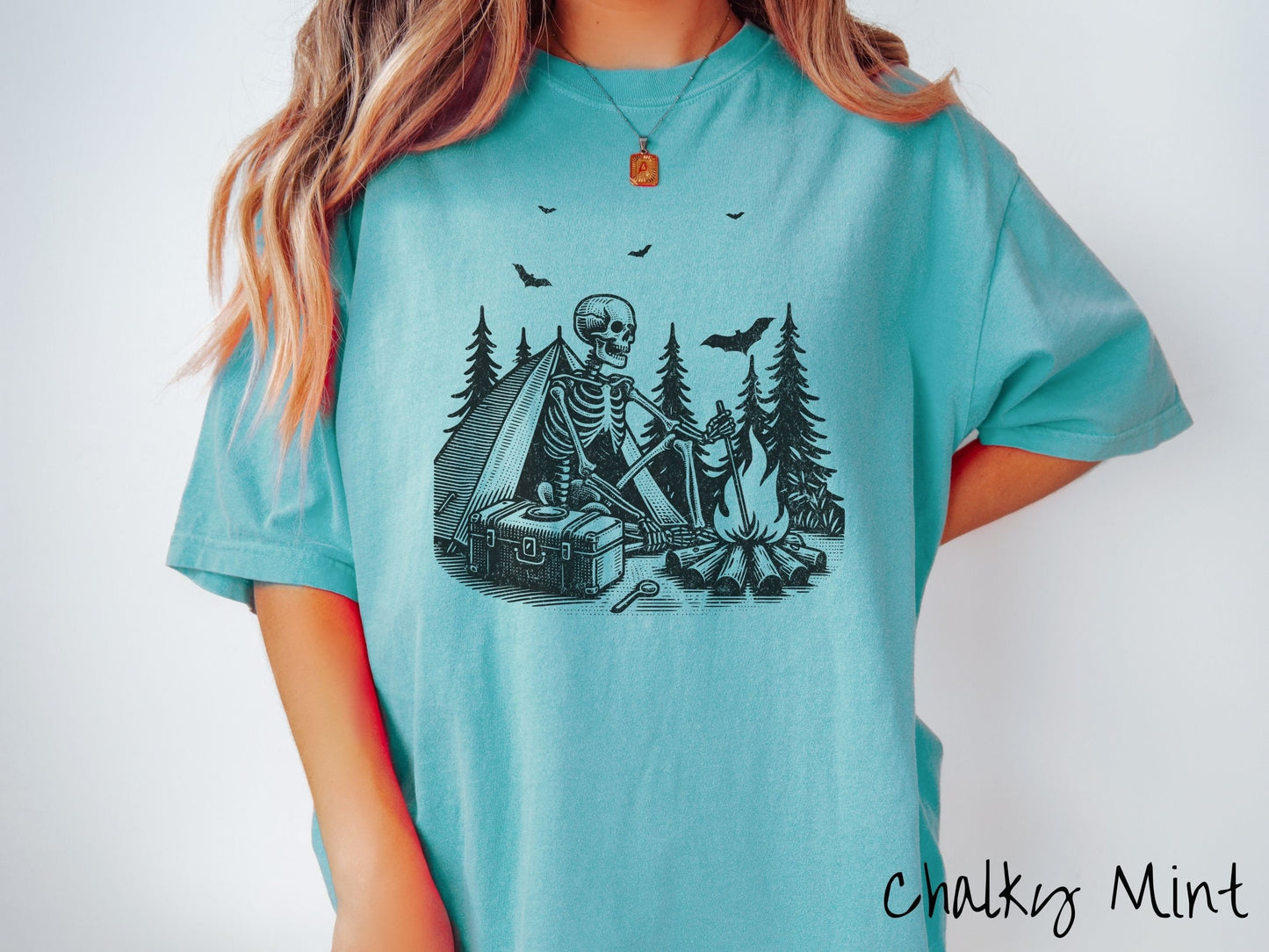 A woman wearing a cute, vintage chalky mint colored Comfort Colors T-shirt with a skeleton sitting in a tent building a fire next to a cooler out in the woods. The campsite is surrounded by evergreen trees and there are bats flying above.
