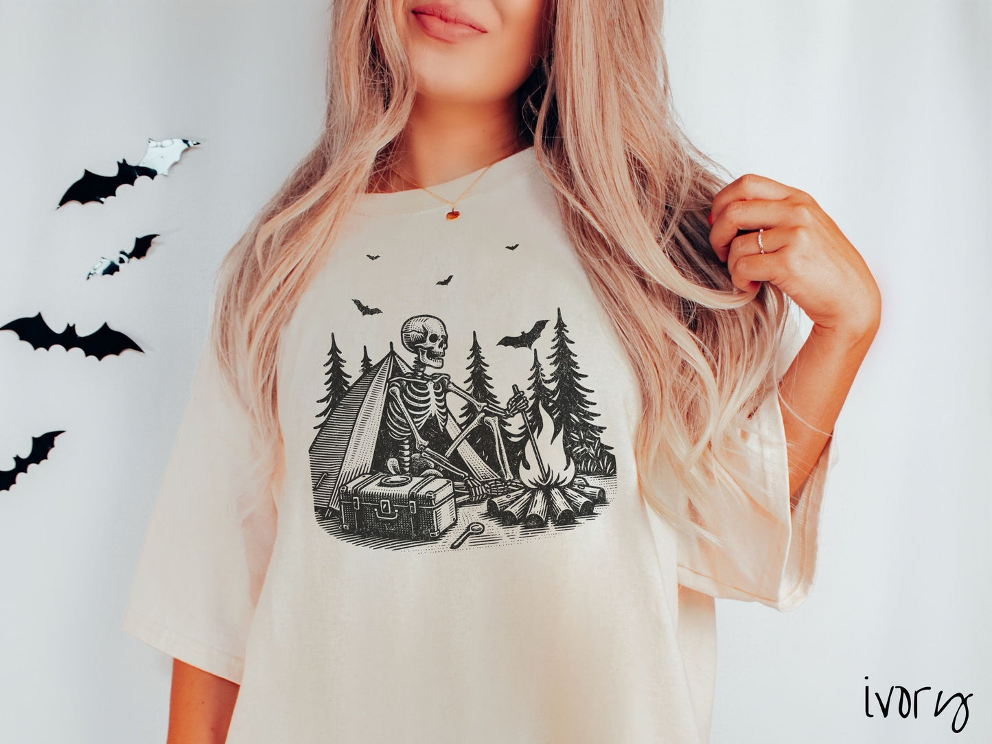 A woman wearing a cute, vintage ivory colored Comfort Colors T-shirt with a skeleton sitting in a tent building a fire next to a cooler out in the woods. The campsite is surrounded by evergreen trees and there are bats flying above.