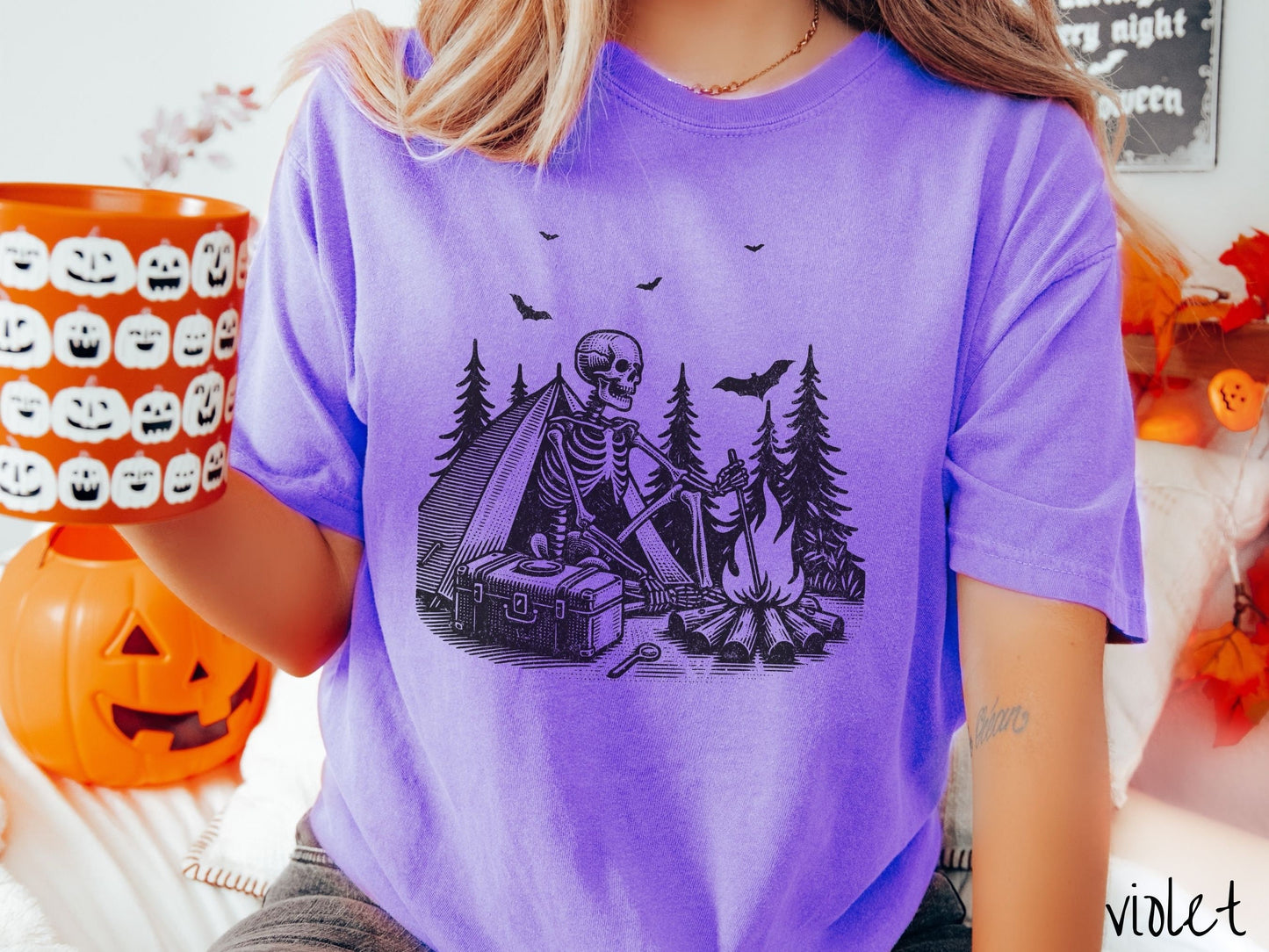 A woman wearing a cute, vintage violet colored Comfort Colors T-shirt with a skeleton sitting in a tent building a fire next to a cooler out in the woods. The campsite is surrounded by evergreen trees and there are bats flying above.