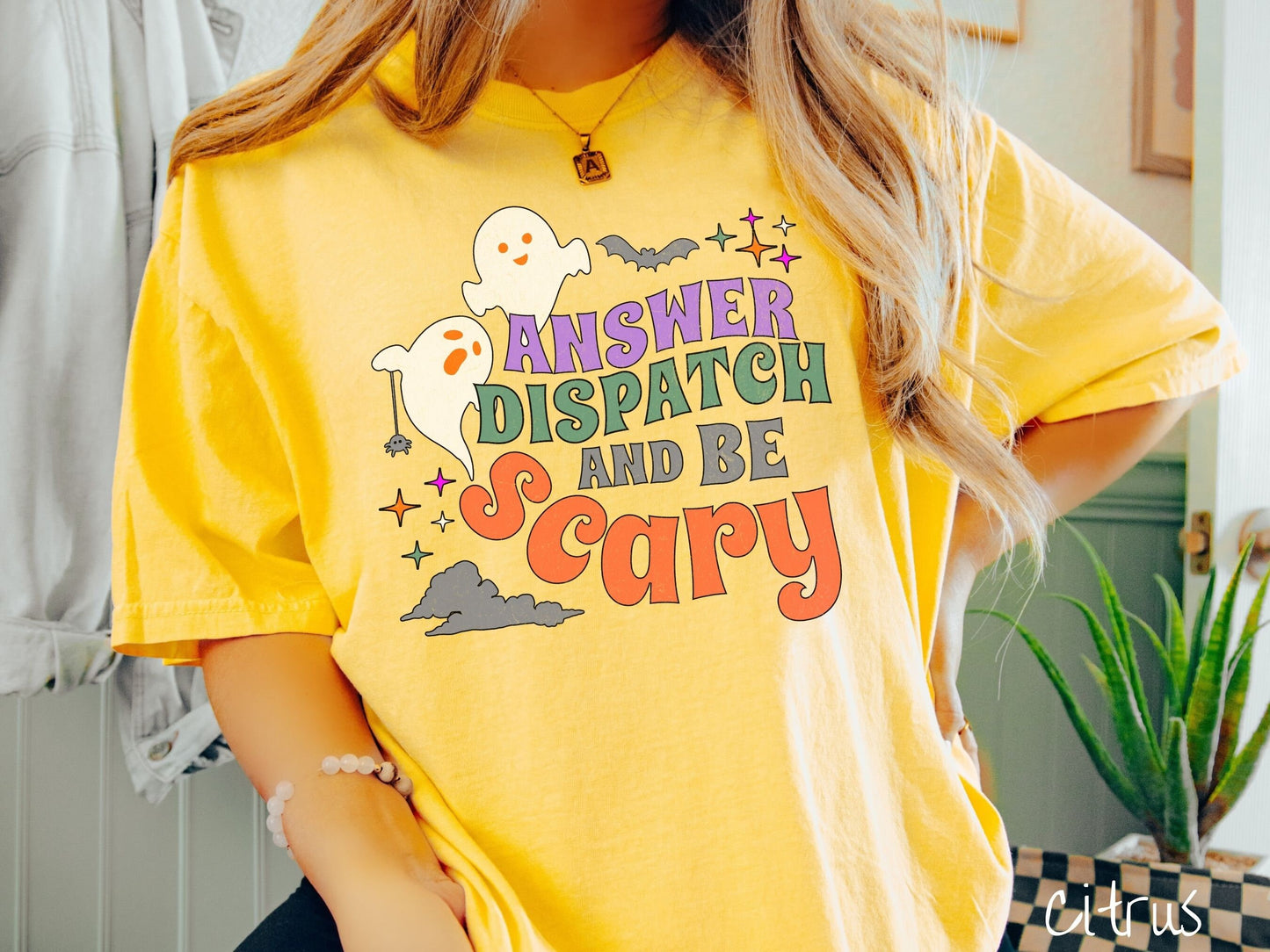 A woman wearing a cute, vintage citrus colored Comfort Colors T-shirt with the text Answer Dispatch and Be scary in colorful font. There are orange-eyed white ghosts flying around and black bats and twinkling stars in the sky.