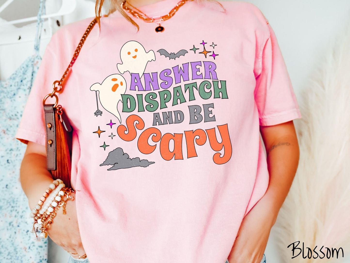 A woman wearing a cute, vintage blossom colored Comfort Colors T-shirt with the text Answer Dispatch and Be scary in colorful font. There are orange-eyed white ghosts flying around and black bats and twinkling stars in the sky.