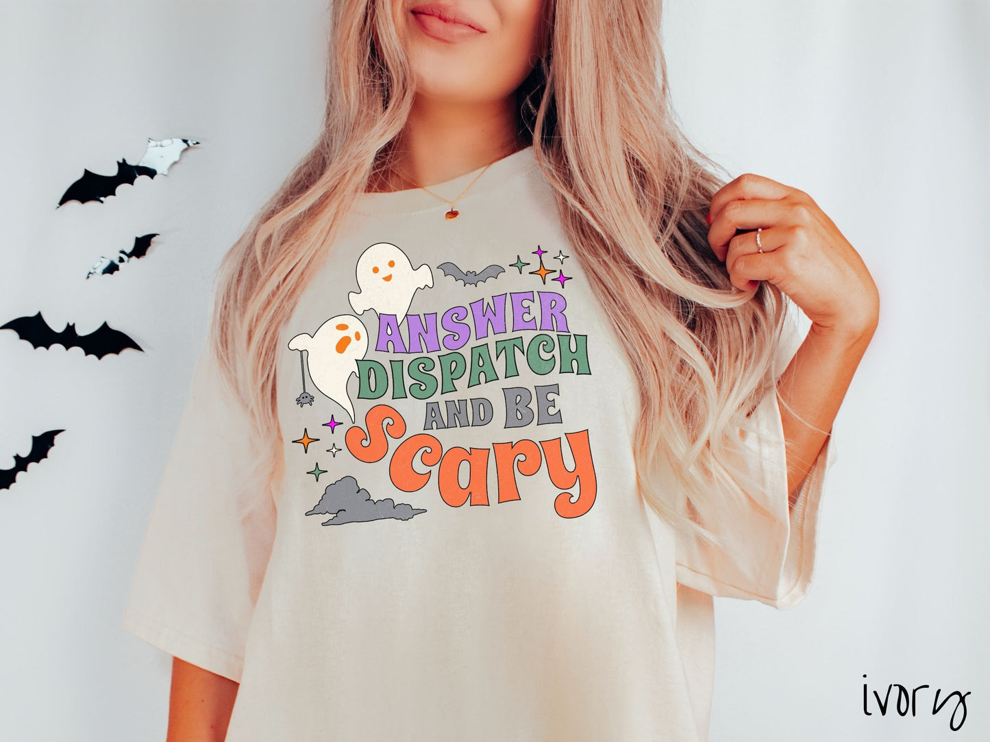 A woman wearing a cute, vintage ivory colored Comfort Colors T-shirt with the text Answer Dispatch and Be scary in colorful font. There are orange-eyed white ghosts flying around and black bats and twinkling stars in the sky.