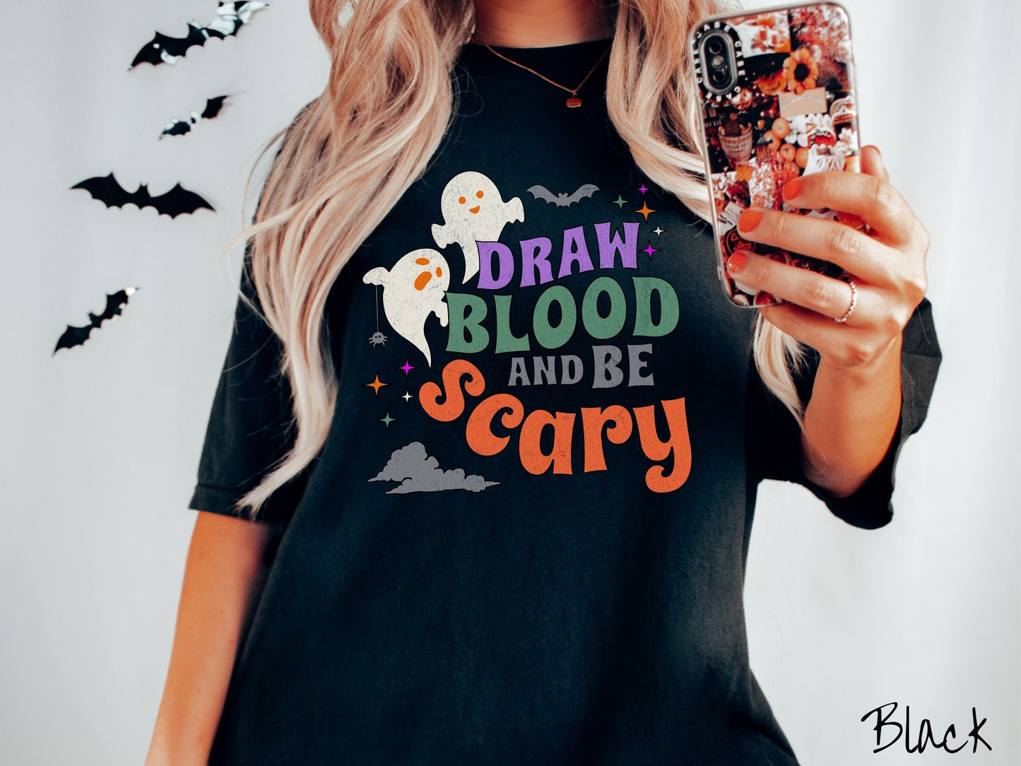 A woman wearing a cute, vintage black colored Comfort Colors T-shirt with the text Draw Blood and Be Scary in colorful font. There are orange-eyed white ghosts flying around and black bats and twinkling stars in the sky.