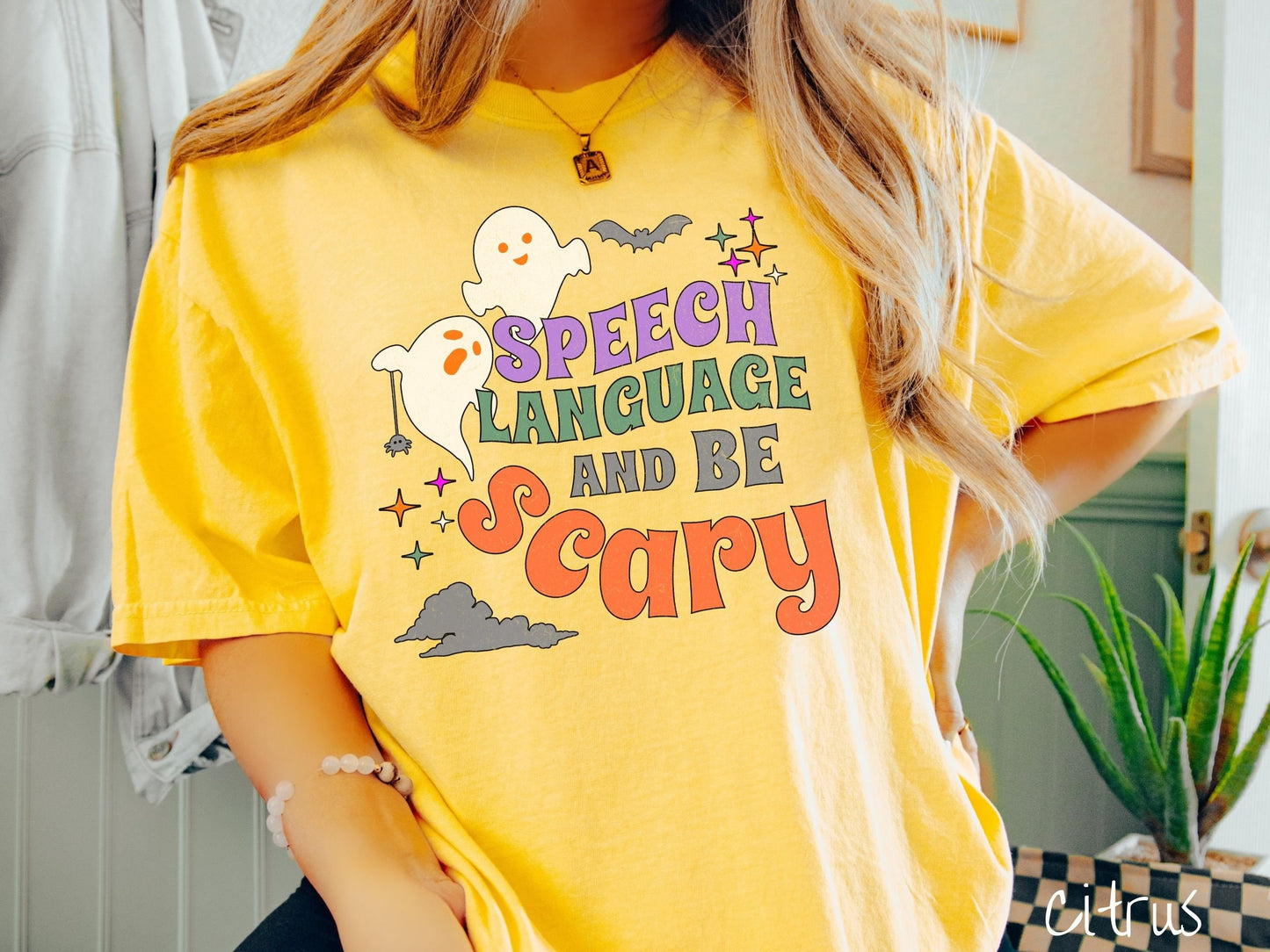 A woman wearing a cute, vintage citrus colored Comfort Colors T-shirt with the text Speech Language and Be Scary in colorful font. There are orange-eyed white ghosts flying around and black bats and twinkling stars in the sky.