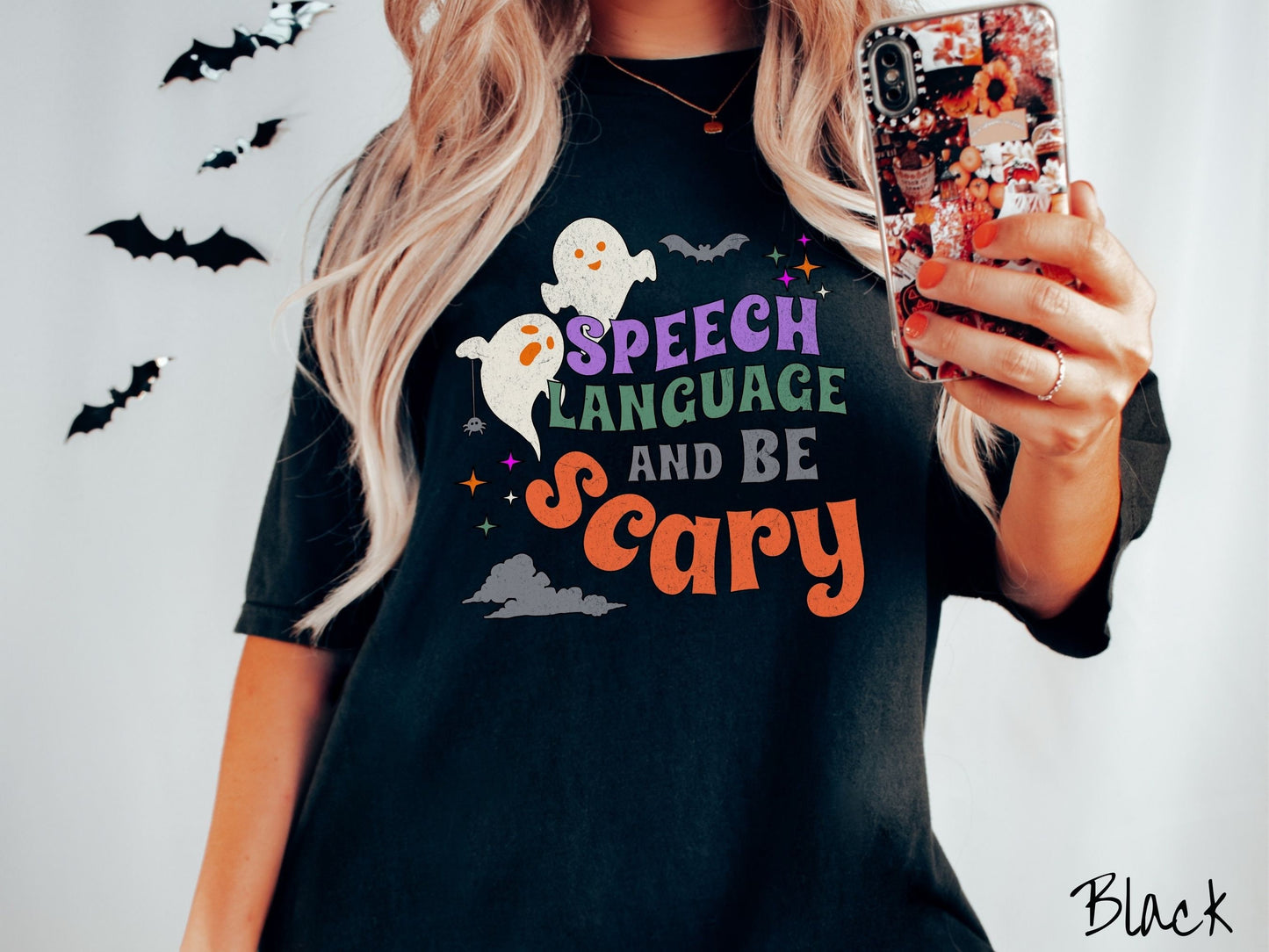A woman wearing a cute, vintage black colored Comfort Colors T-shirt with the text Speech Language and Be Scary in colorful font. There are orange-eyed white ghosts flying around and black bats and twinkling stars in the sky.
