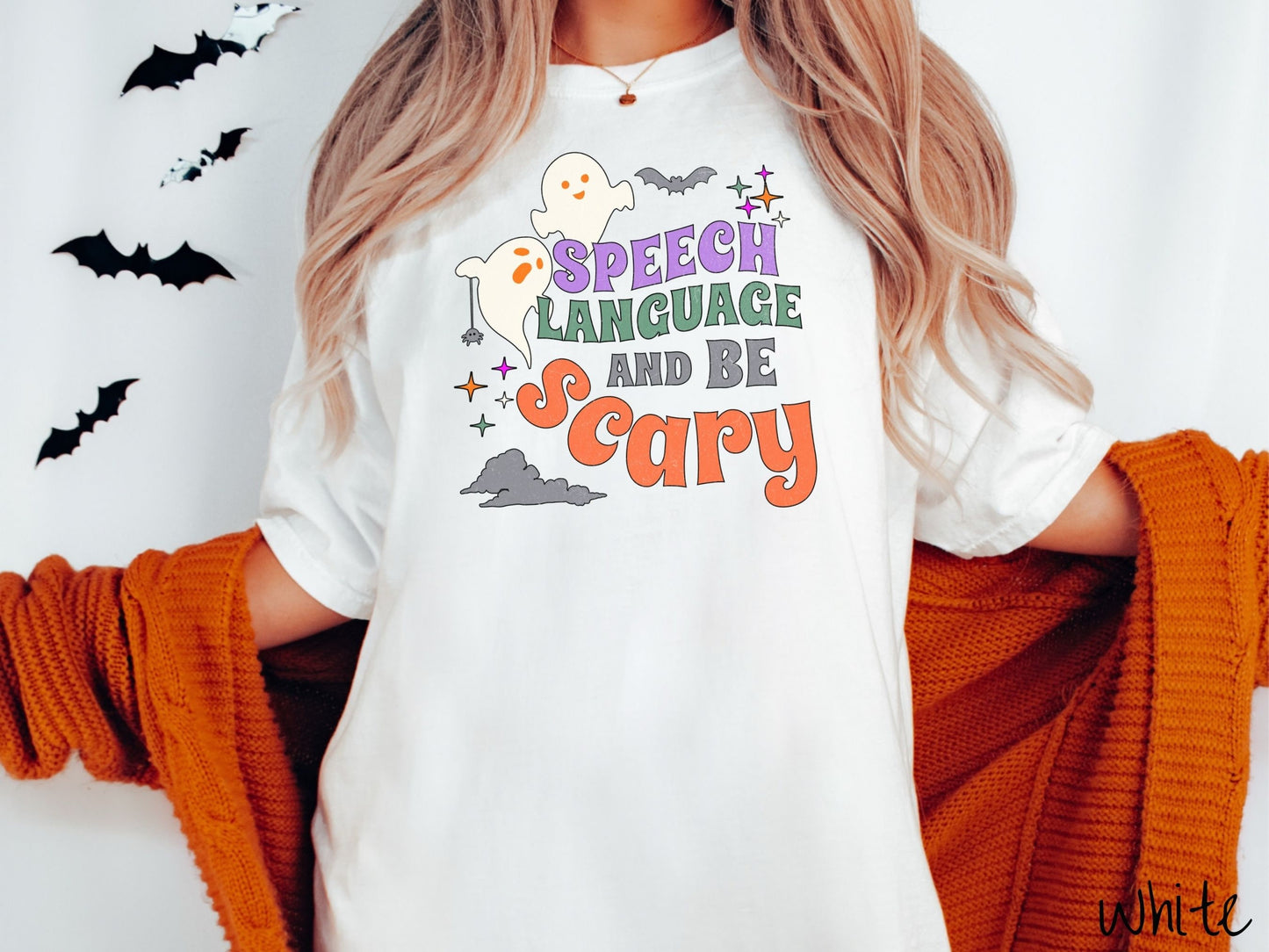 A woman wearing a cute, vintage white colored Comfort Colors T-shirt with the text Speech Language and Be Scary in colorful font. There are orange-eyed white ghosts flying around and black bats and twinkling stars in the sky.