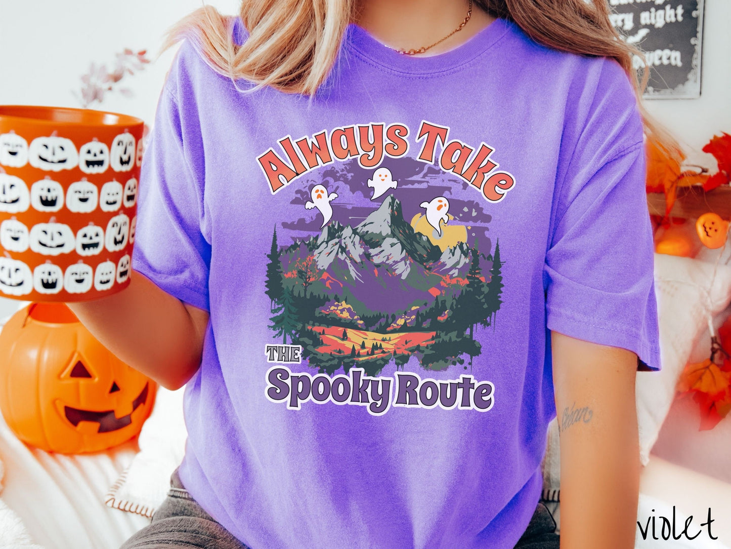 A woman wearing a cute, vintage violet colored Comfort Colors T-shirt with the text Always Take the Spooky Route in colorful font. There are three white ghosts with orange faces flying above a mountain landscape with rolling hills and green forest.