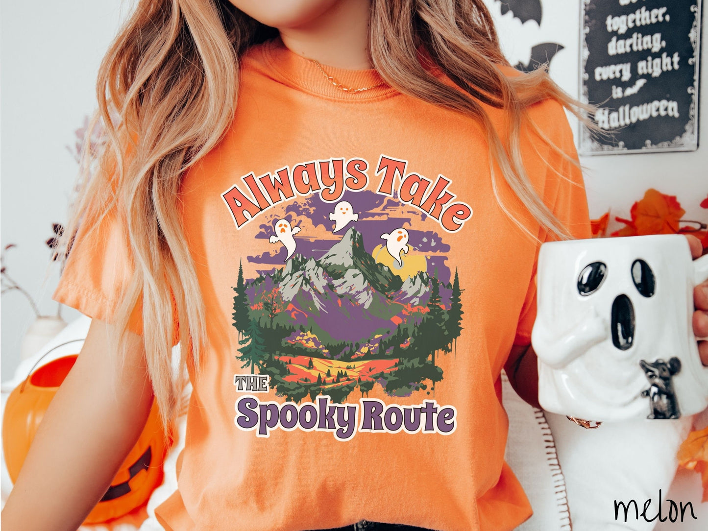 A woman wearing a cute, vintage melon colored Comfort Colors T-shirt with the text Always Take the Spooky Route in colorful font. There are three white ghosts with orange faces flying above a mountain landscape with rolling hills and green forest.