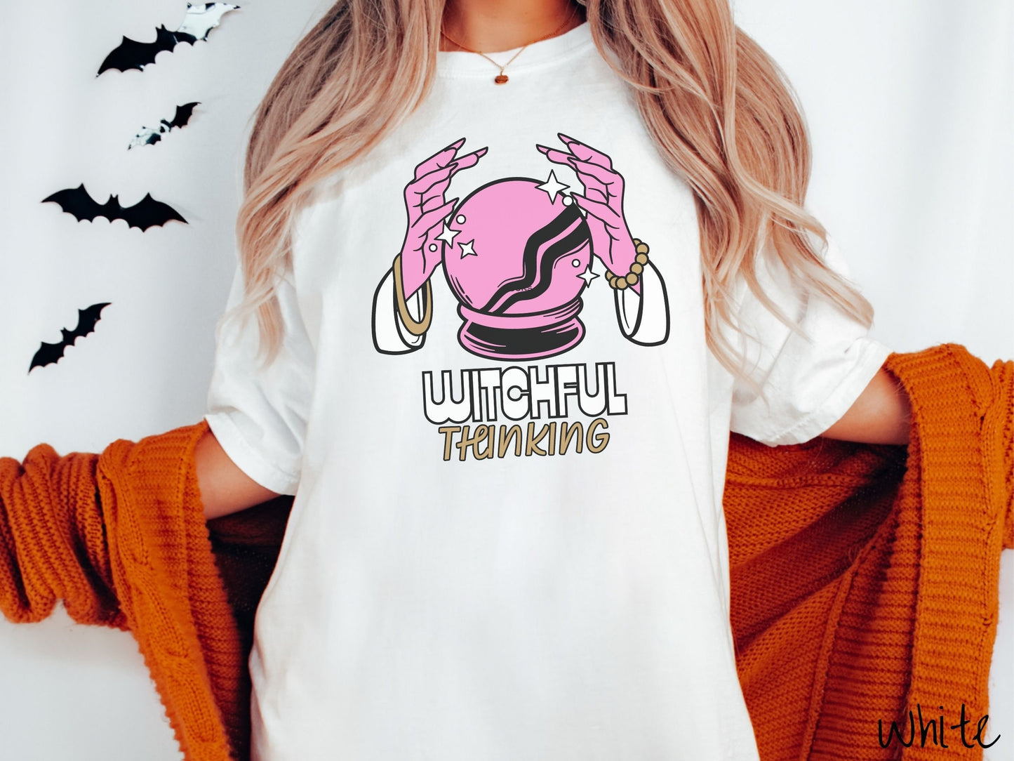 A woman wearing a cute, vintage white colored Comfort Colors T-shirt with the text Witchful Thinking in white and gold font. Above is a pink crystal ball, and there are two pink witch hands with jewelry hovering around the crystall ball.