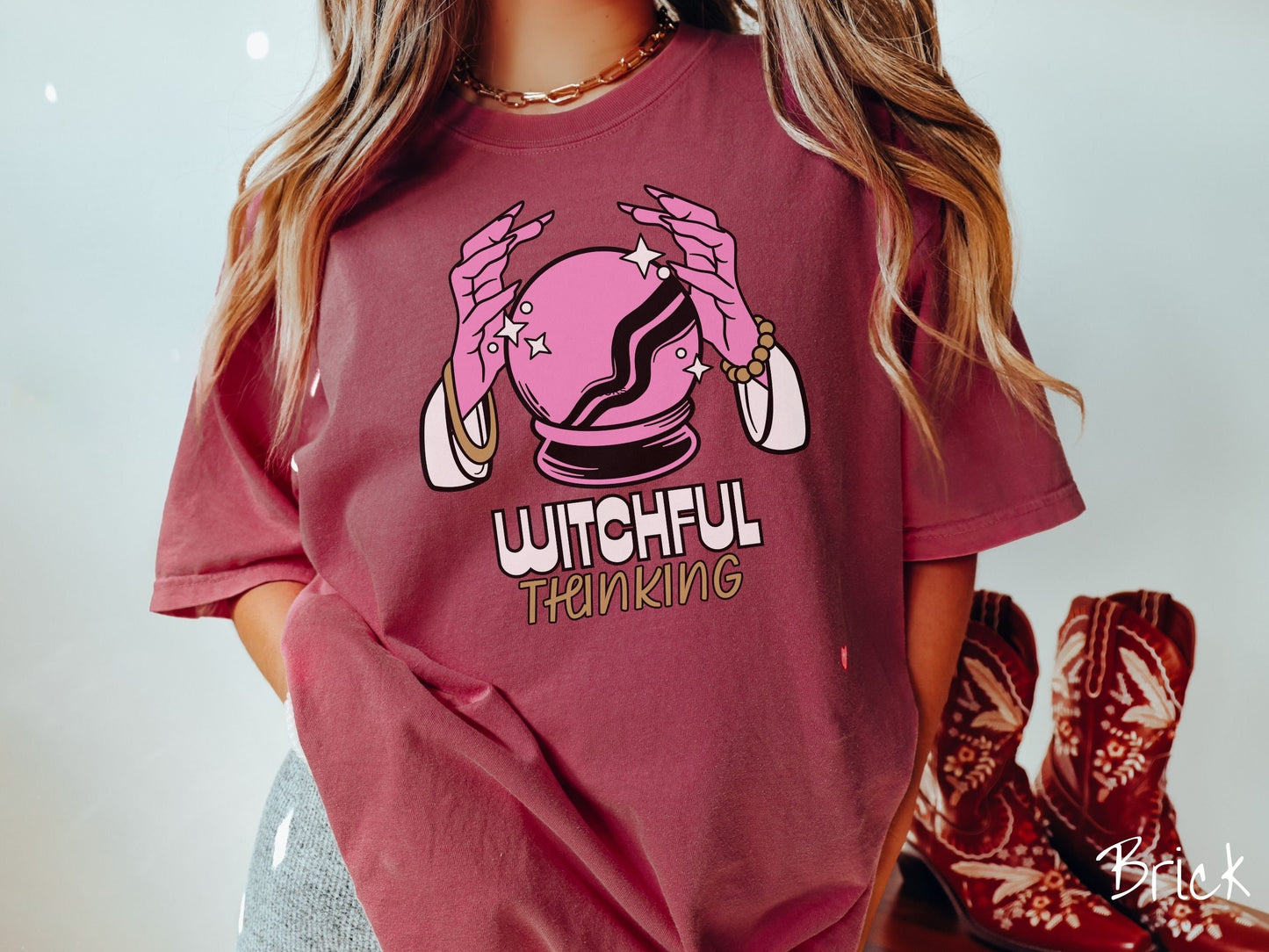 A woman wearing a cute, vintage brick colored Comfort Colors T-shirt with the text Witchful Thinking in white and gold font. Above is a pink crystal ball, and there are two pink witch hands with jewelry hovering around the crystall ball.