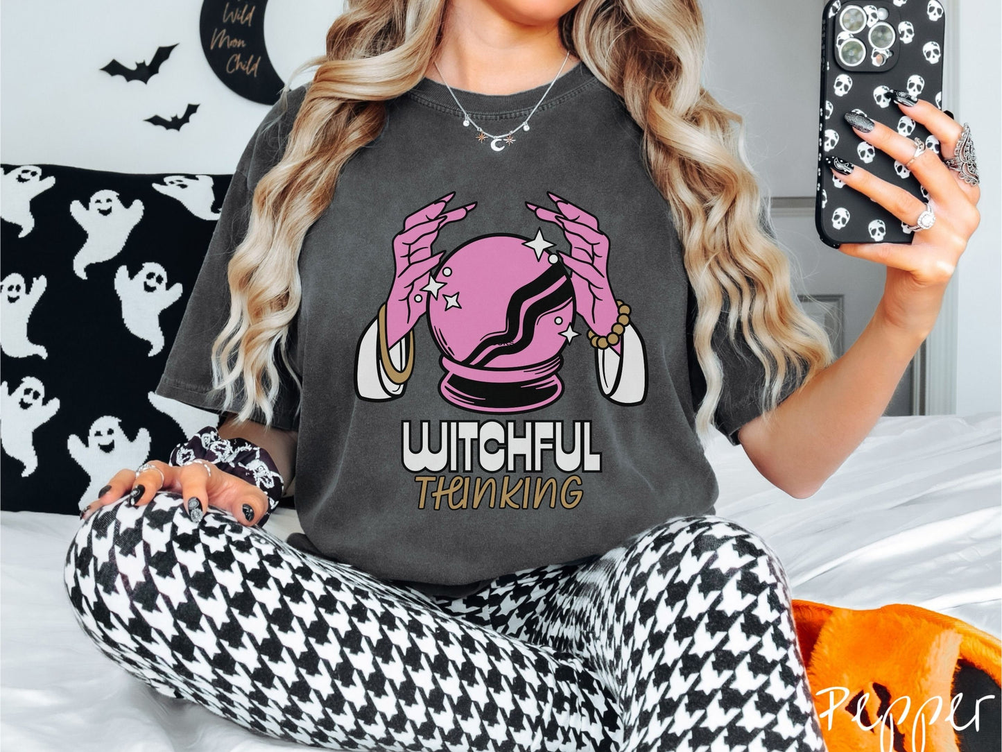 A woman wearing a cute, vintage pepper colored Comfort Colors T-shirt with the text Witchful Thinking in white and gold font. Above is a pink crystal ball, and there are two pink witch hands with jewelry hovering around the crystall ball.