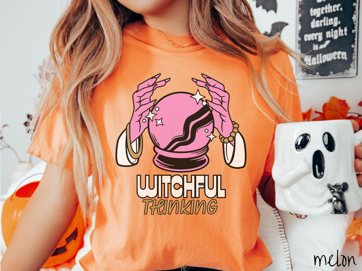 A woman wearing a cute, vintage melon colored Comfort Colors T-shirt with the text Witchful Thinking in white and gold font. Above is a pink crystal ball, and there are two pink witch hands with jewelry hovering around the crystall ball.