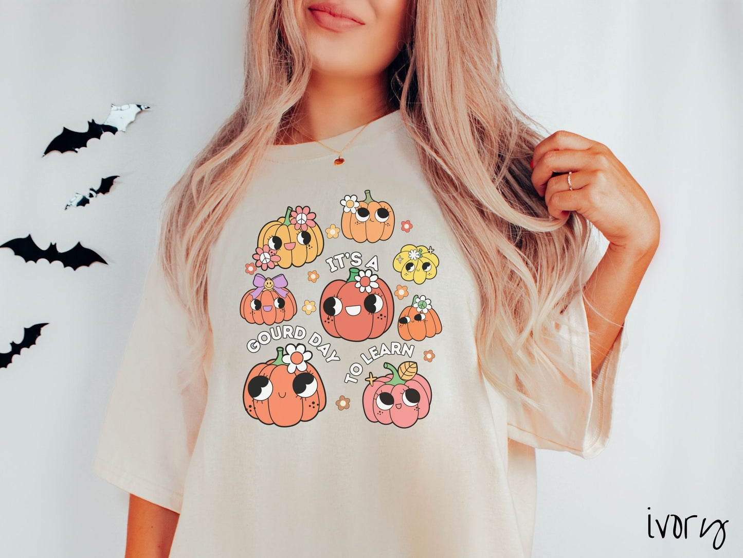A woman wearing a cute, vintage ivory colored Comfort Colors T-shirt with the text Its a Gourd Day to Learn in white font. There is a three by three grid of orange, yellow, and pink pumpkins with girly eyes, wearing flowers and bows.