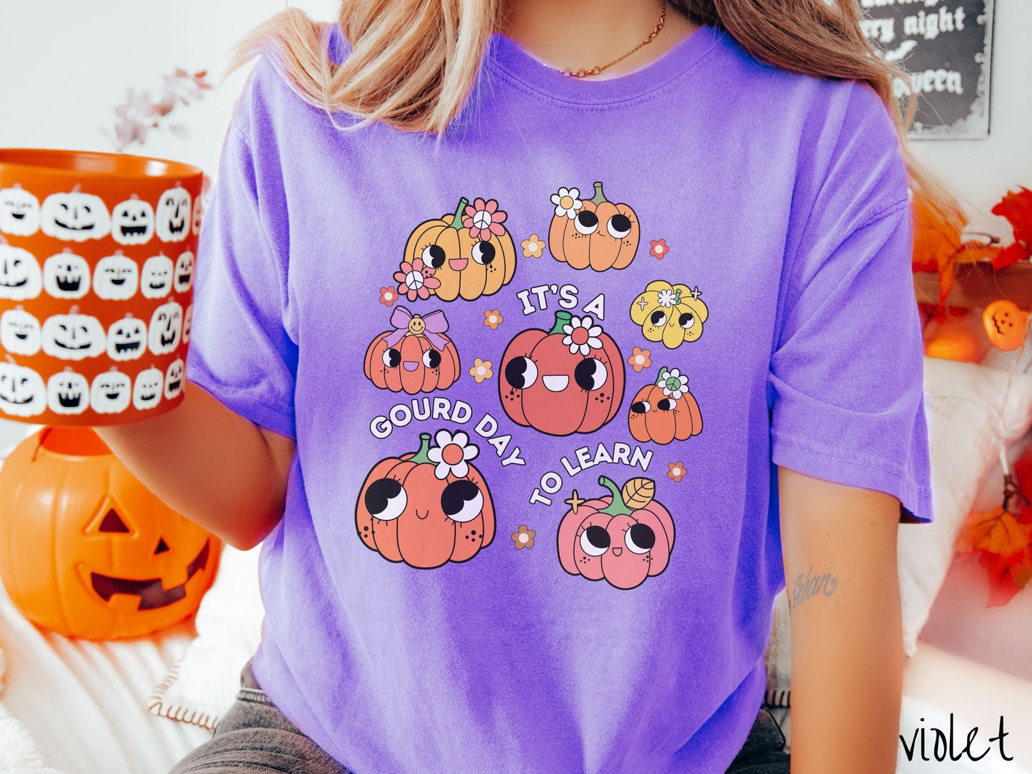 A woman wearing a cute, vintage violet colored Comfort Colors T-shirt with the text Its a Gourd Day to Learn in white font. There is a three by three grid of orange, yellow, and pink pumpkins with girly eyes, wearing flowers and bows.