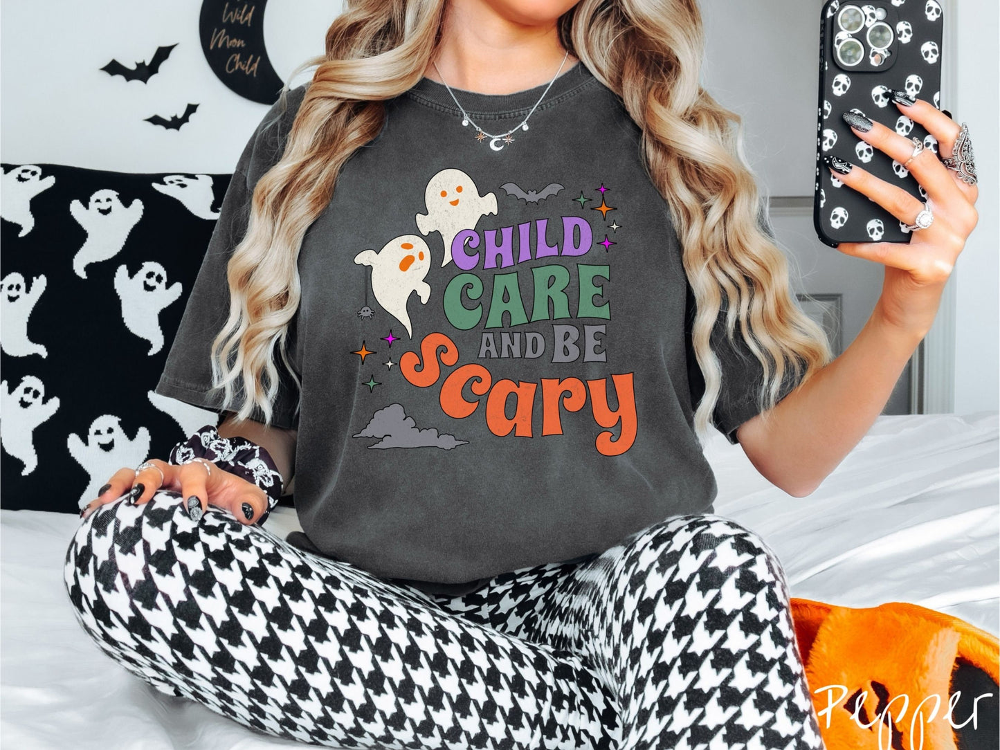 A woman wearing a cute, vintage pepper colored Comfort Colors T-shirt with the text Child Care and Be Scary in colorful font. There are orange-eyed white ghosts flying around and black bats and twinkling stars in the sky.