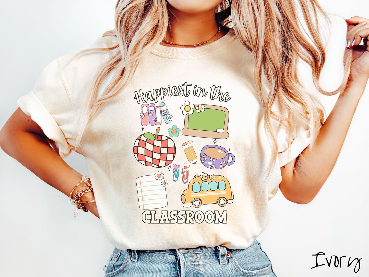 A woman wearing a cute, vintage ivory colored Comfort Colors T-shirt with the text Happiest in the Classroom. In between the text are books, an apple, chalkboard, pencil and paper, coffee cup, yellow school bus, paperclips, and flowers.