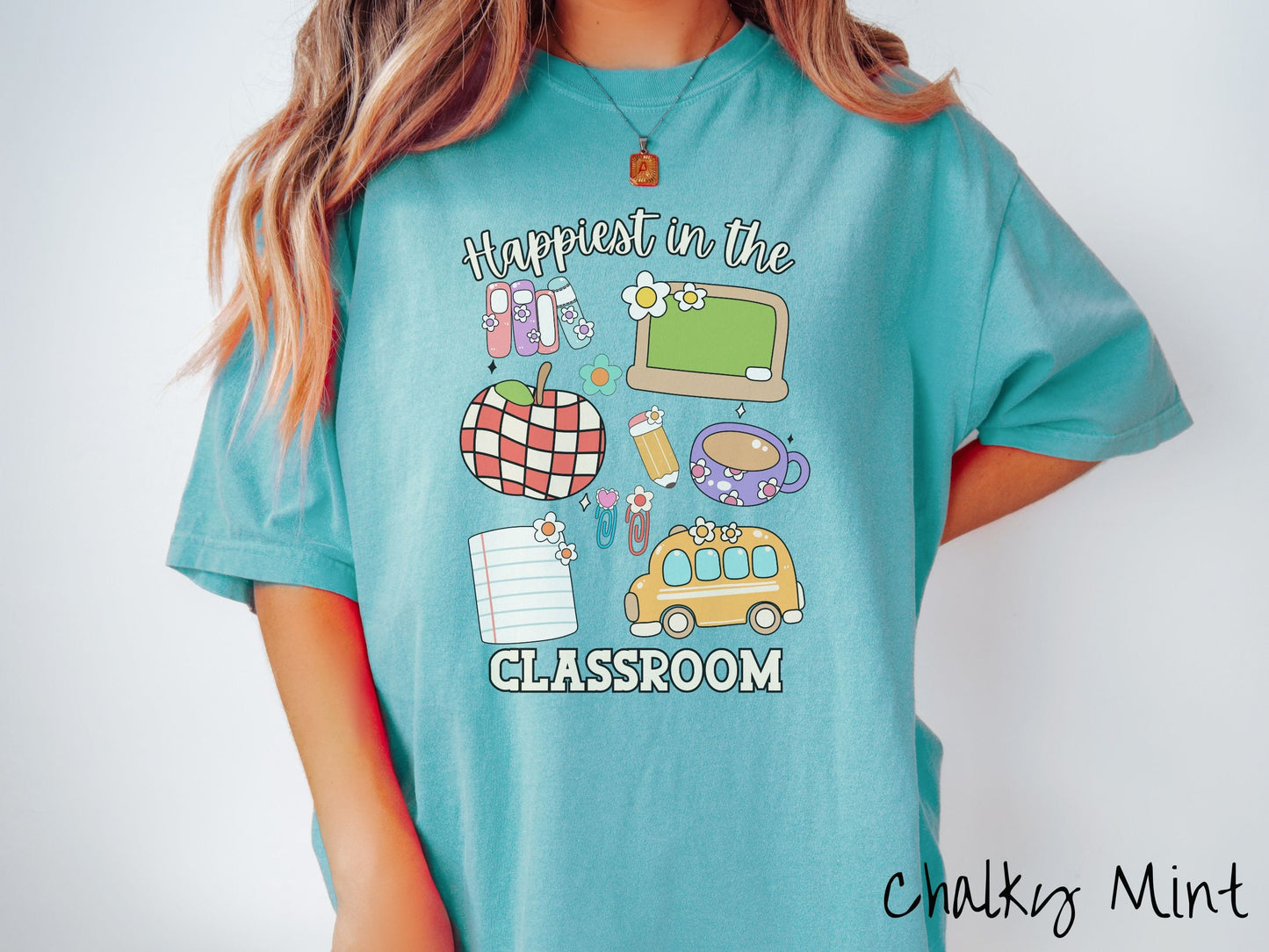 A woman wearing a cute, vintage chalky mint colored Comfort Colors T-shirt with the text Happiest in the Classroom. In between the text are books, an apple, chalkboard, pencil and paper, coffee cup, yellow school bus, paperclips, and flowers.