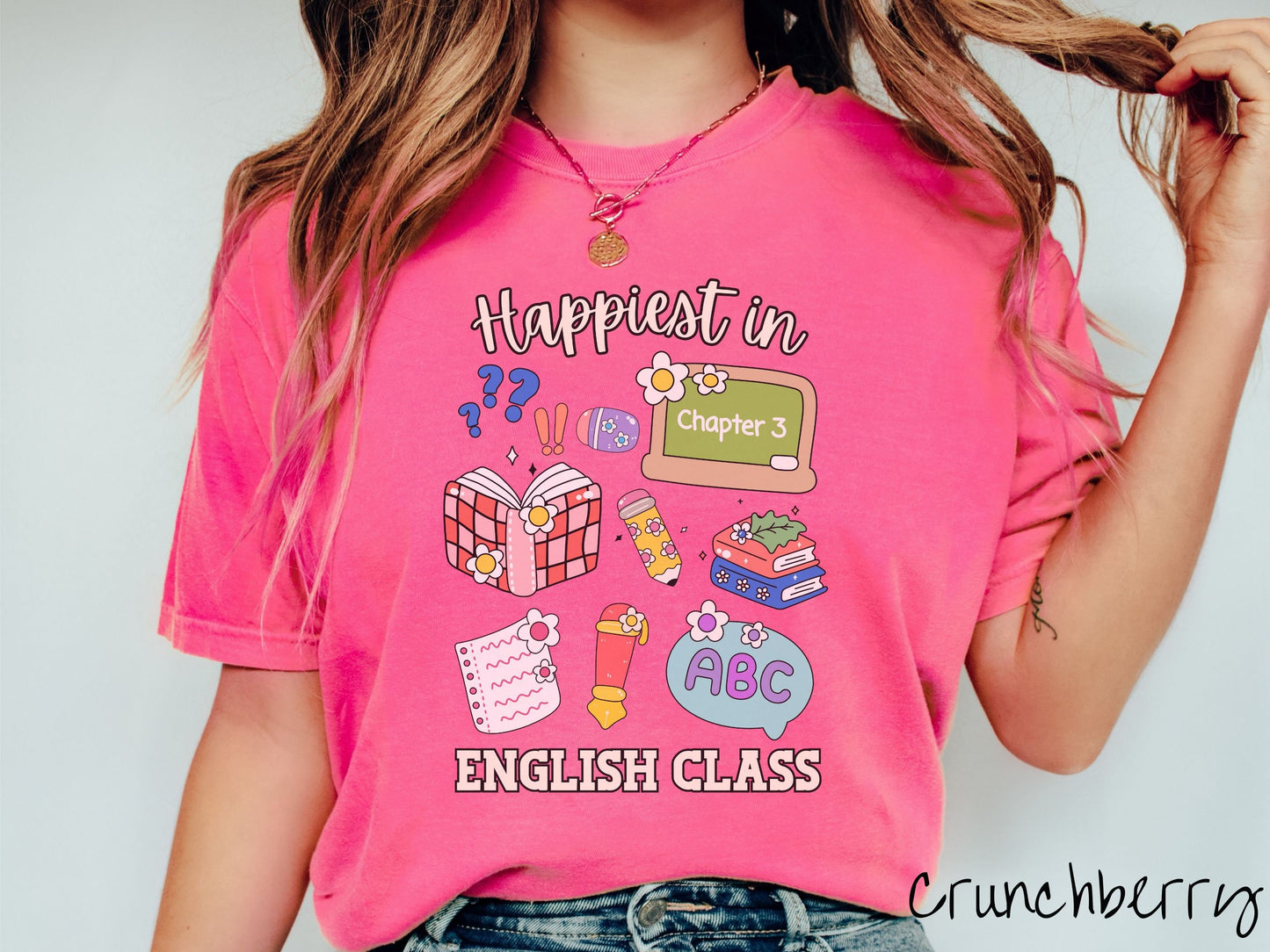 A woman wearing a cute, vintage crunchberry colored Comfort Colors T-shirt with the text Happiest in English Class. In between the text are books, erasers, a chalkboard, pencil, pen, notebook paper, alphabet letters, and flowers.