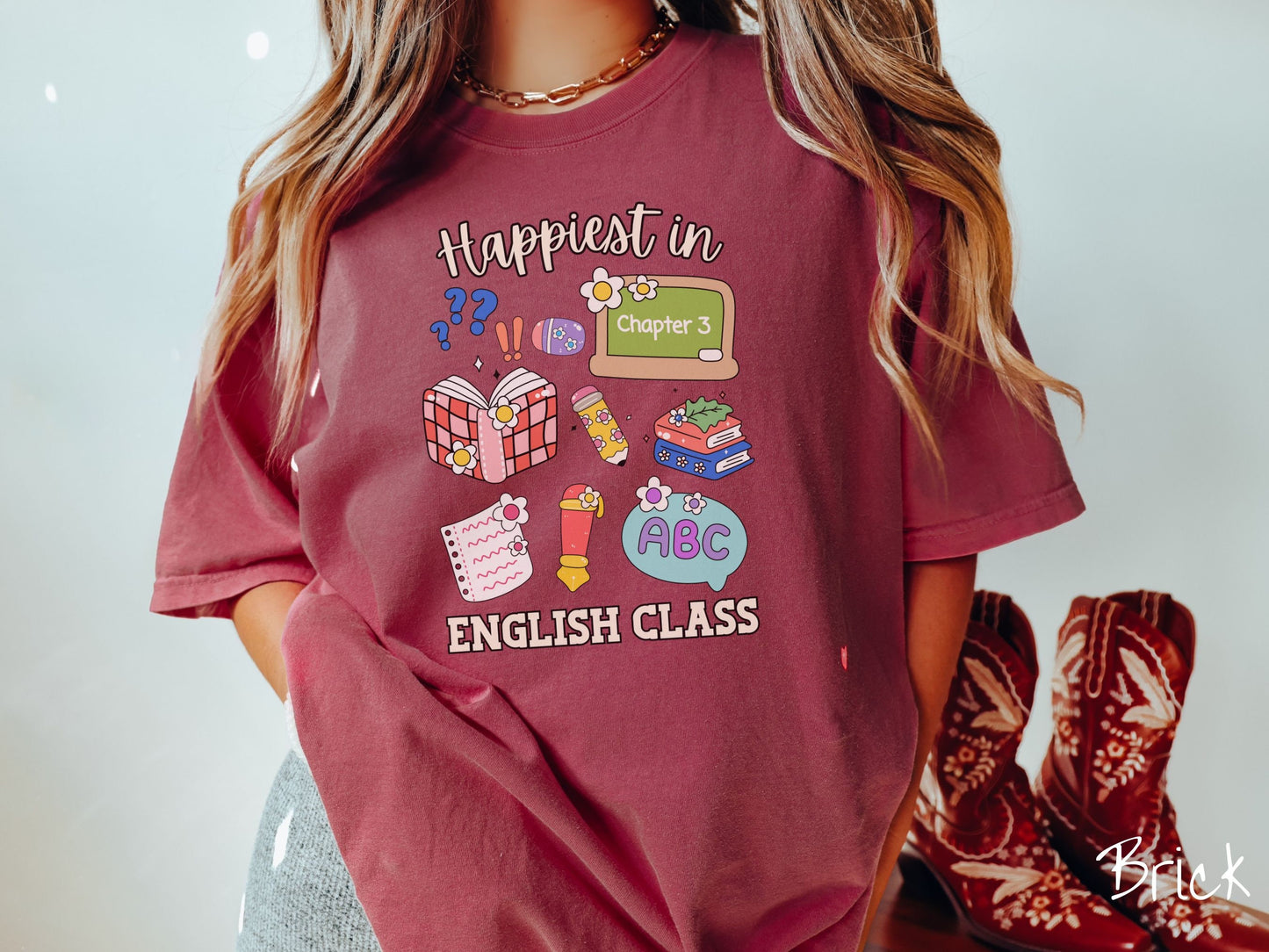 A woman wearing a cute, vintage brick colored Comfort Colors T-shirt with the text Happiest in English Class. In between the text are books, erasers, a chalkboard, pencil, pen, notebook paper, alphabet letters, and flowers.