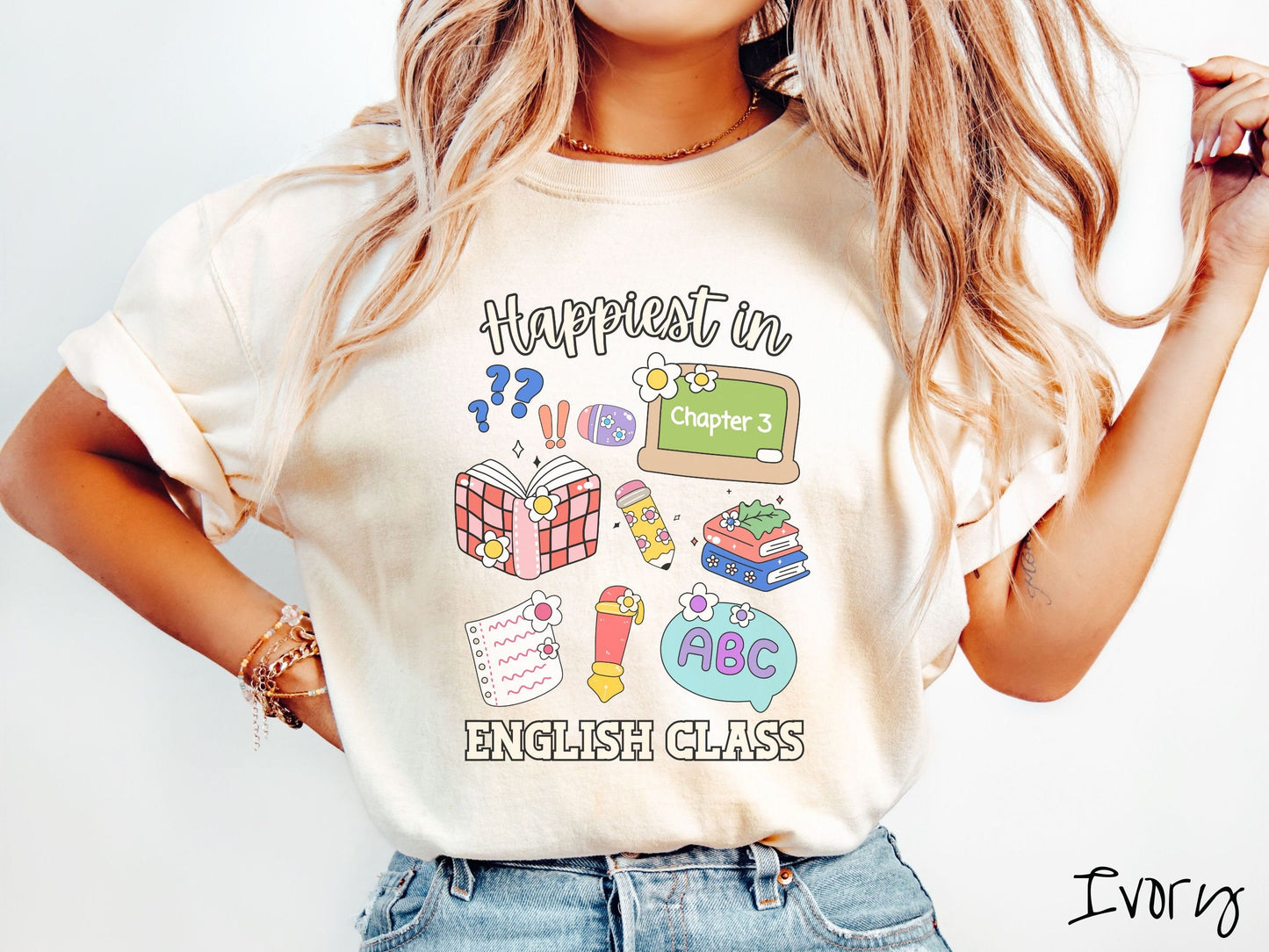 A woman wearing a cute, vintage ivory colored Comfort Colors T-shirt with the text Happiest in English Class. In between the text are books, erasers, a chalkboard, pencil, pen, notebook paper, alphabet letters, and flowers.