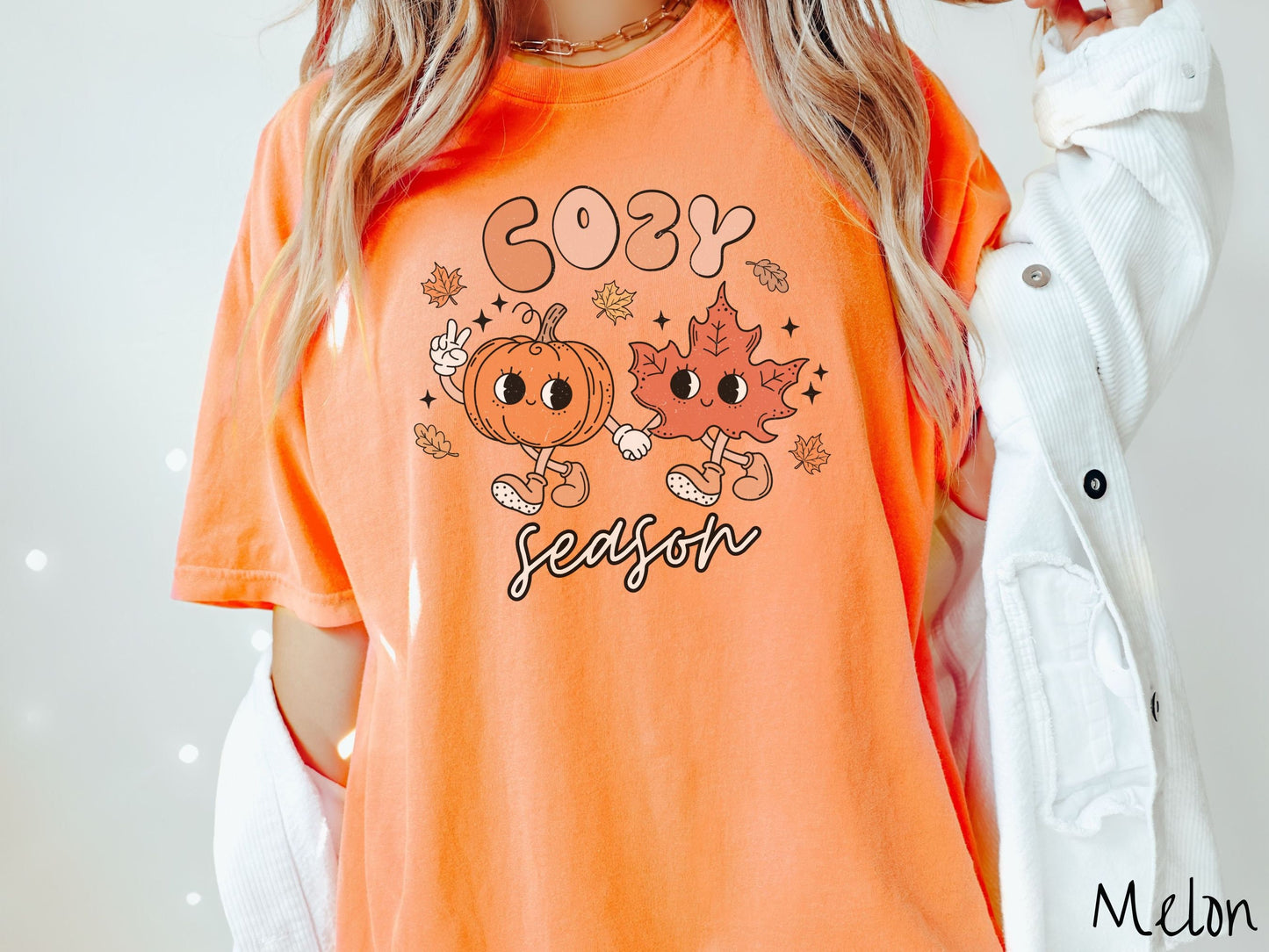 A woman wearing a cute, vintage melon colored Comfort Colors T-shirt with the text Cozy Season and in between the text are a smiling orange pumpkin holding up a peace sign with one hand and holding hands with a smiling dark orange leaf.