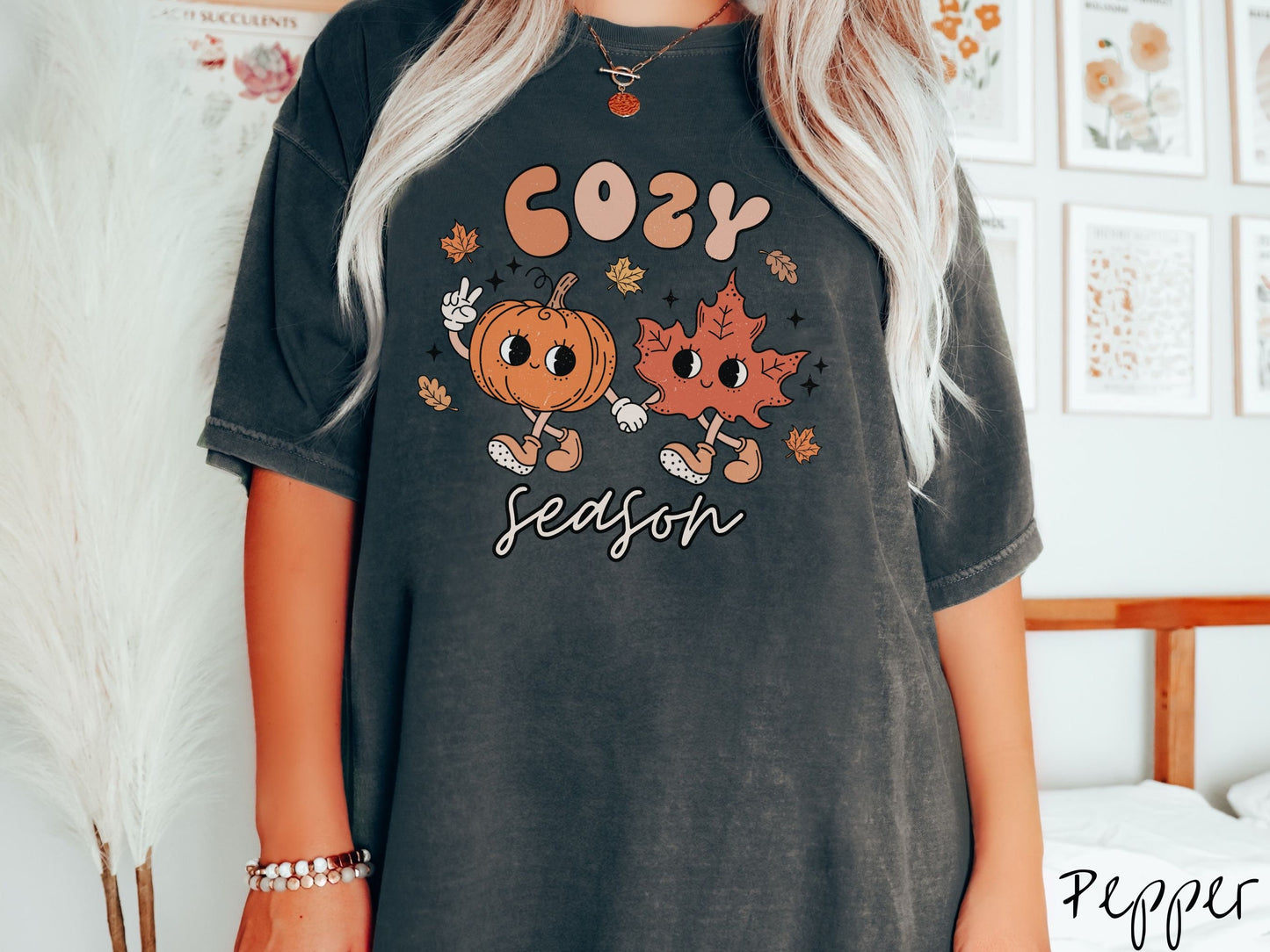A woman wearing a cute, vintage pepper colored Comfort Colors T-shirt with the text Cozy Season and in between the text are a smiling orange pumpkin holding up a peace sign with one hand and holding hands with a smiling dark orange leaf.