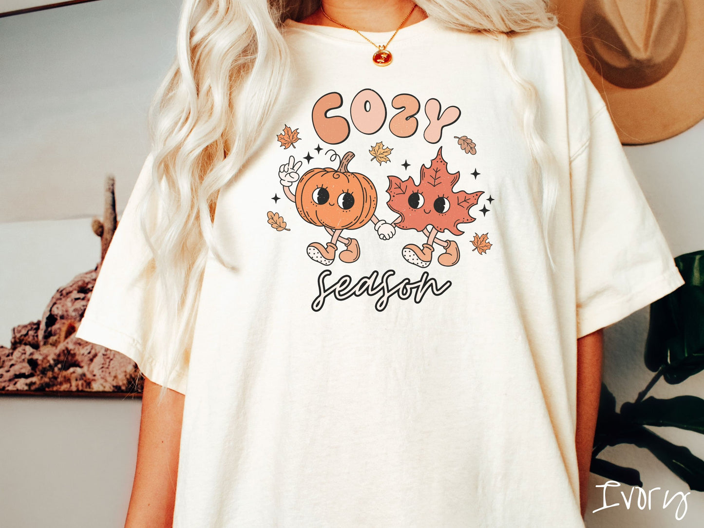 A woman wearing a cute, vintage ivory colored Comfort Colors T-shirt with the text Cozy Season and in between the text are a smiling orange pumpkin holding up a peace sign with one hand and holding hands with a smiling dark orange leaf.