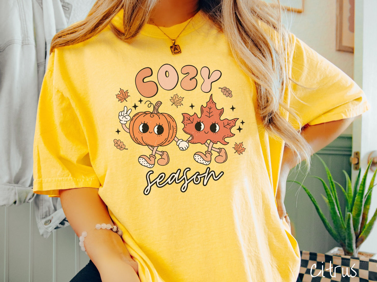 A woman wearing a cute, vintage citrus colored Comfort Colors T-shirt with the text Cozy Season and in between the text are a smiling orange pumpkin holding up a peace sign with one hand and holding hands with a smiling dark orange leaf.