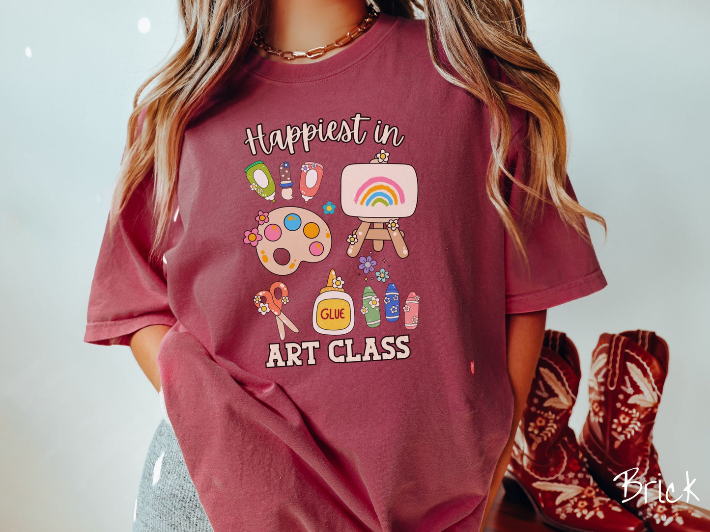 A woman wearing a cute, vintage brick colored Comfort Colors T-shirt with the text Happiest in Art Class. In between the text are paint brushes, a paint palette, an art canvas on a stand, scissors, glue, crayons, and flowers.