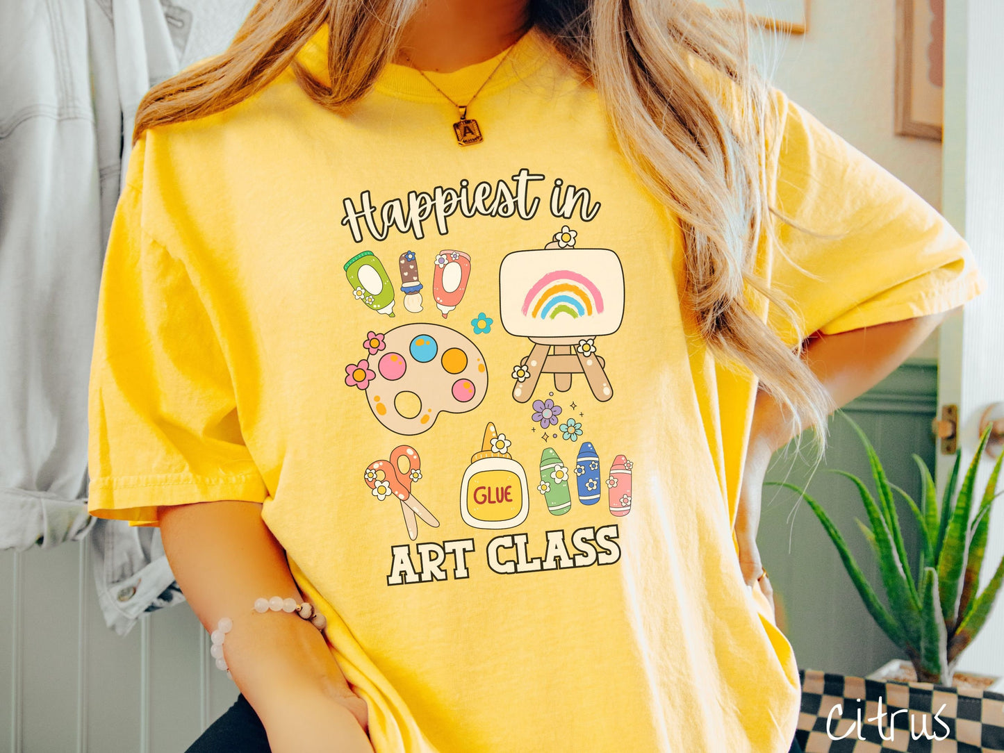 A woman wearing a cute, vintage citrus colored Comfort Colors T-shirt with the text Happiest in Art Class. In between the text are paint brushes, a paint palette, an art canvas on a stand, scissors, glue, crayons, and flowers.