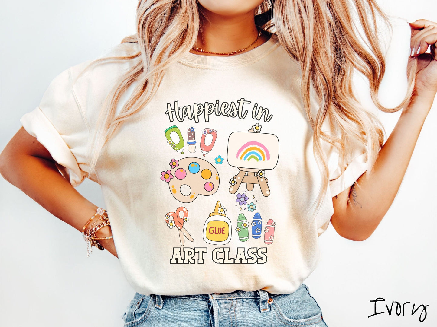 A woman wearing a cute, vintage ivory colored Comfort Colors T-shirt with the text Happiest in Art Class. In between the text are paint brushes, a paint palette, an art canvas on a stand, scissors, glue, crayons, and flowers.