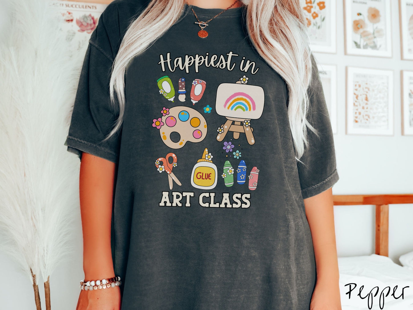 A woman wearing a cute, vintage pepper colored Comfort Colors T-shirt with the text Happiest in Art Class. In between the text are paint brushes, a paint palette, an art canvas on a stand, scissors, glue, crayons, and flowers.