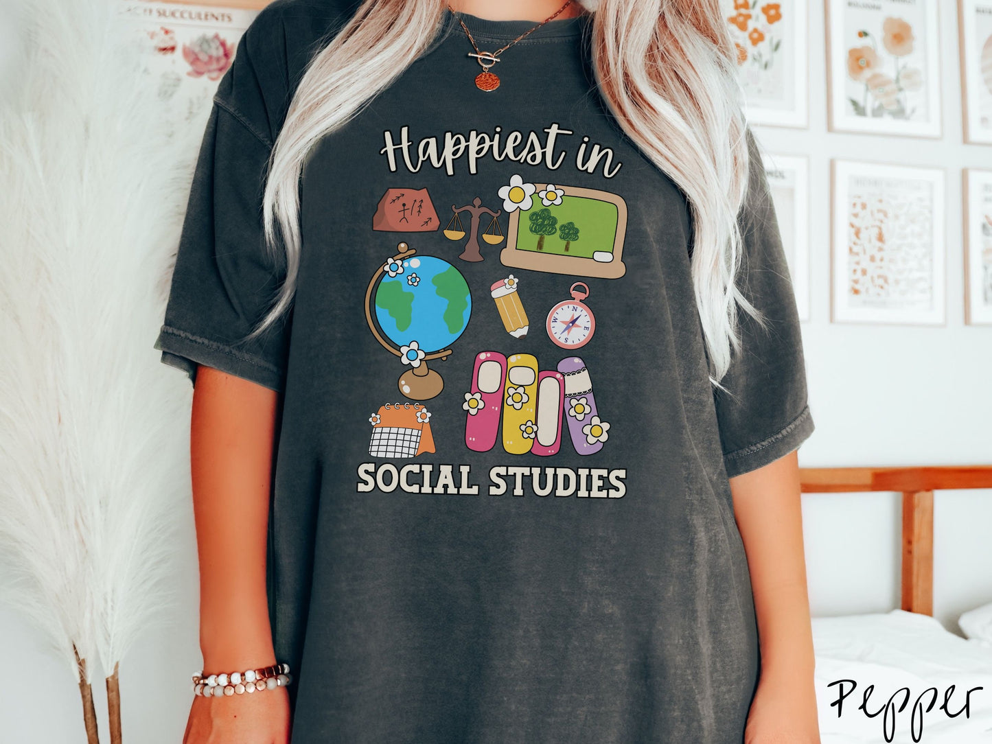 A woman wearing a cute, vintage pepper colored Comfort Colors T-shirt with the text Happiest in Social Studies Class. In between the text are hieroglyphics, a globe, a pencil, compass, books, laptop computer, and a weight scale.