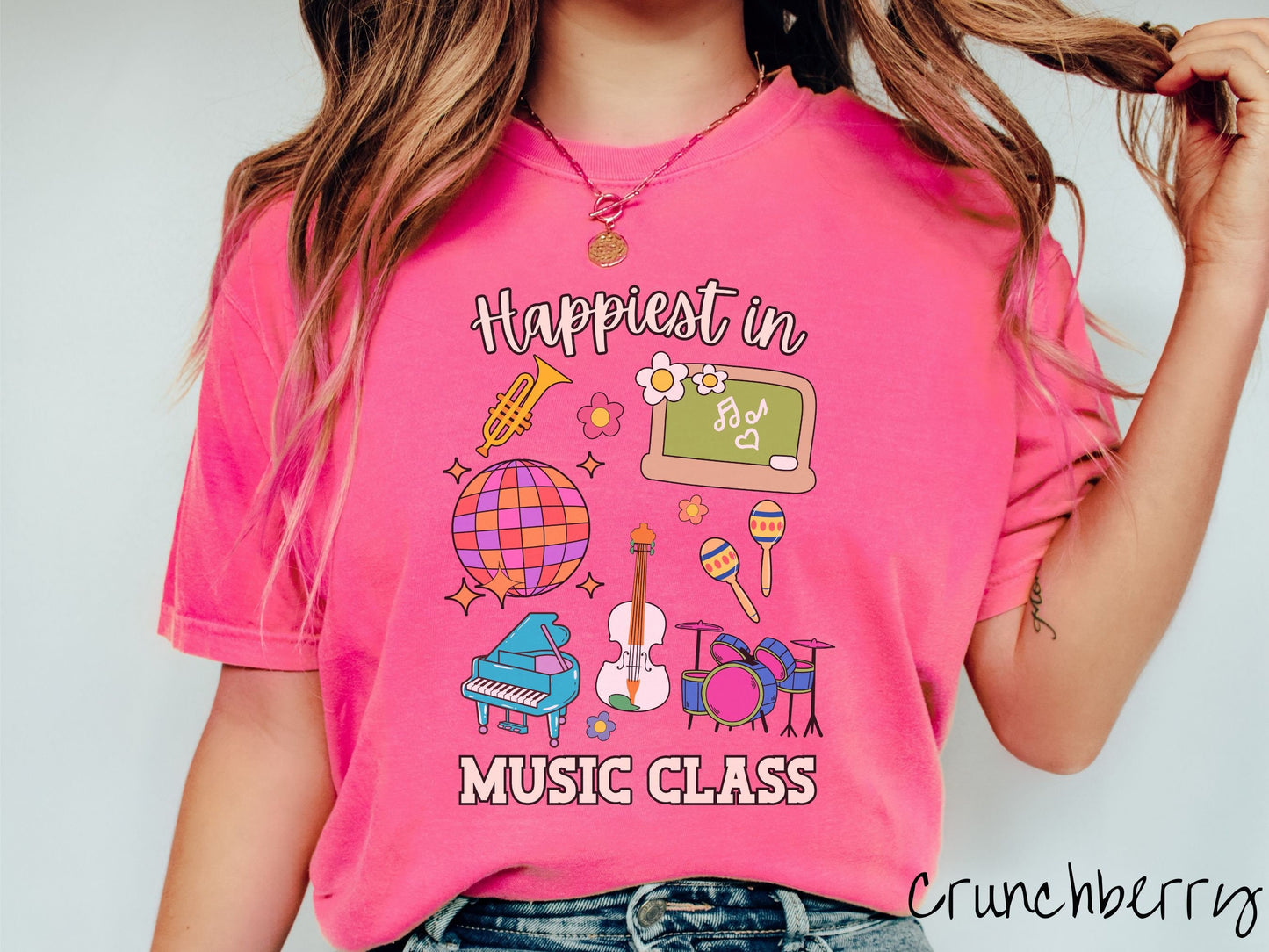 A woman wearing a cute, vintage crunchberry colored Comfort Colors T-shirt with the text Happiest in Music Class. In between the text are a saxophone, a disco ball, a chalkboard, a piano, a guitar, a drum set, maracas, and flowers.