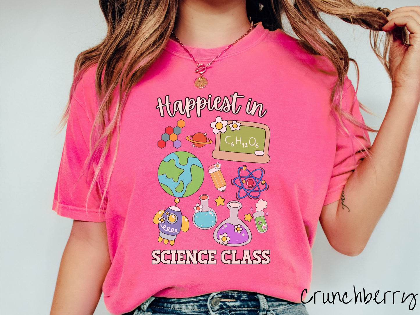 A woman wearing a cute, vintage crunchberry colored Comfort Colors T-shirt with the text Happiest in Science Class. In between the text are a globe, chemistry, rocket ship, chalkboard, science test tubes, a pencil, DNA molecules, and Saturn.