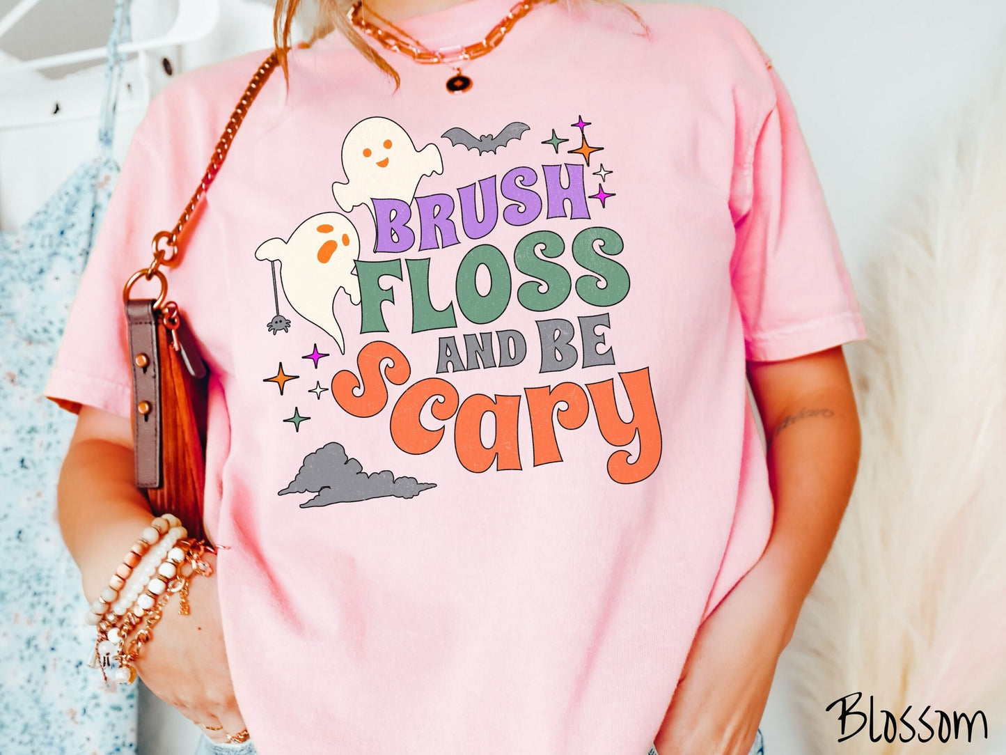 A woman wearing a cute, vintage blossom colored Comfort Colors T-shirt with the text Brush Floss and Be Scary in colorful font. There are orange-eyed white ghosts flying around and black bats and twinkling stars in the sky.