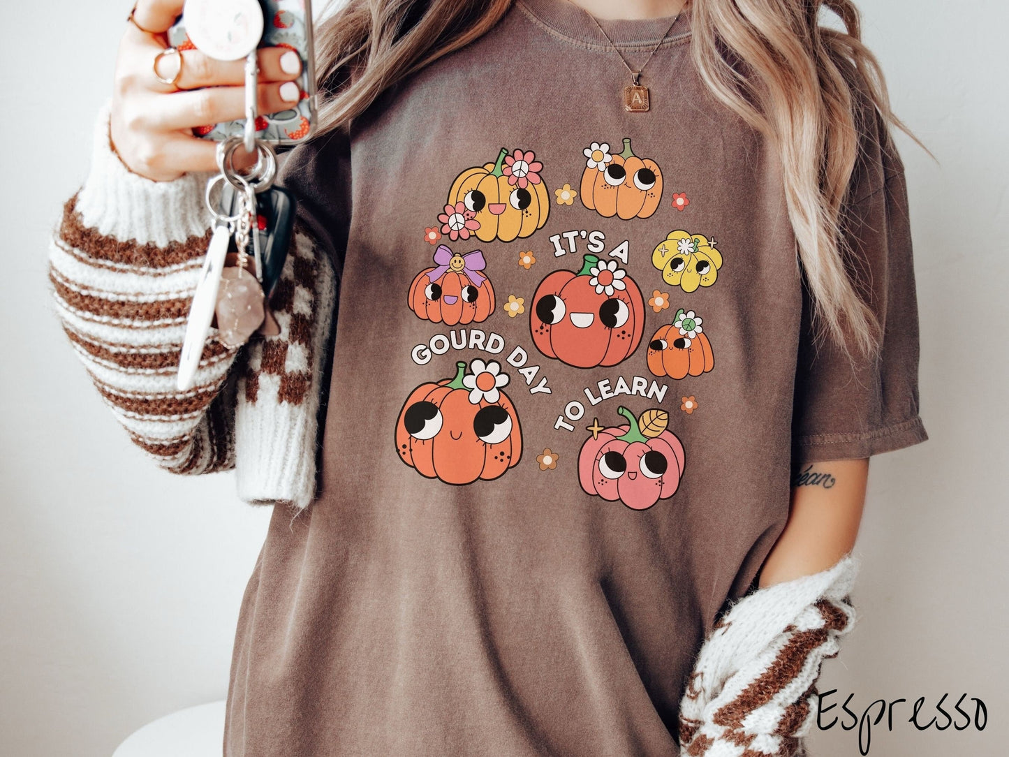A woman wearing a cute, vintage espresso colored Comfort Colors T-shirt with the text Its a Gourd Day to Learn in white font. There is a three by three grid of orange, yellow, and pink pumpkins with girly eyes, wearing flowers and bows.