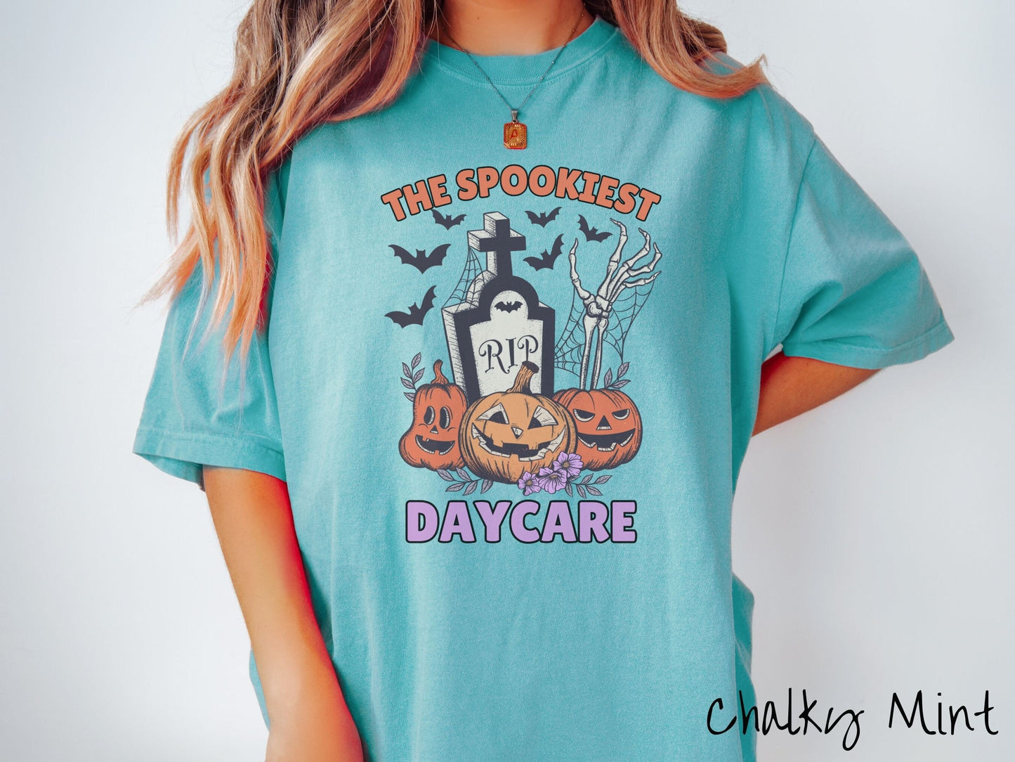A woman wearing a cute, vintage chalky mint colored Comfort Colors T-shirt with text The Spookiest Daycare. There are three carved pumpkins sitting in front of a tombstone with a skeleton arm coming out of the ground and black bats in the air.