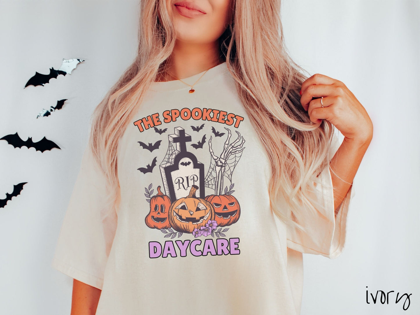 A woman wearing a cute, vintage ivory colored Comfort Colors T-shirt with the text The Spookiest Daycare. There are three smiling carved pumpkins sitting in front of a tombstone with a skeleton arm coming out of the ground and black bats in the air.