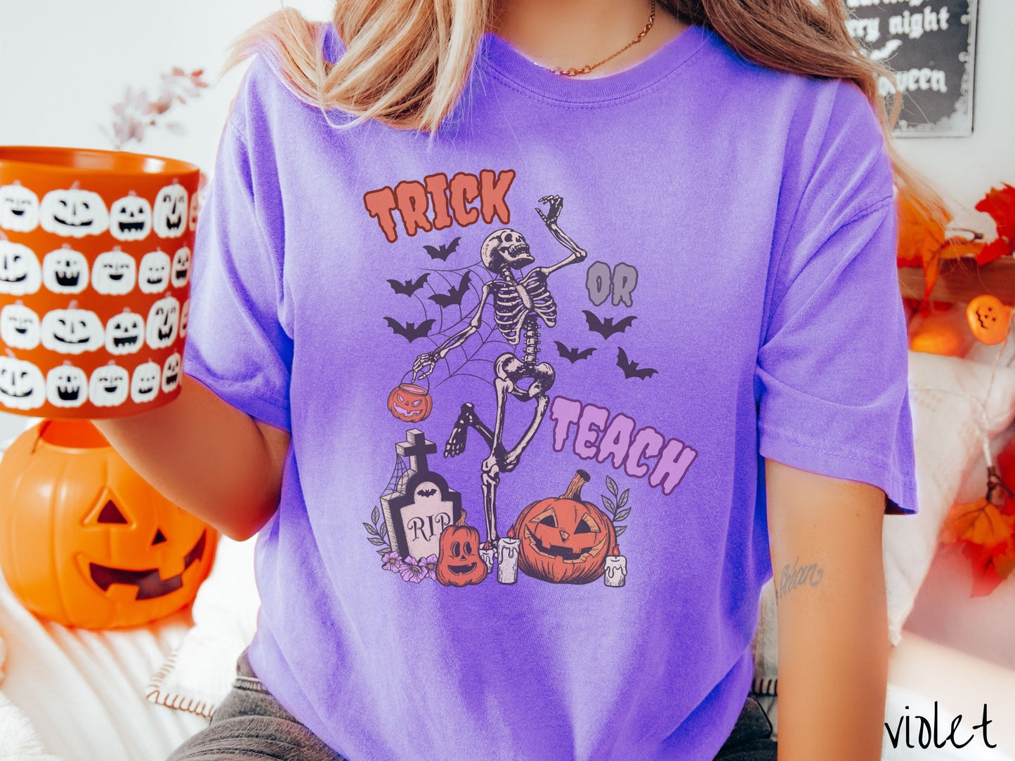 A woman wearing a cute, vintage violet colored Comfort Colors T-shirt with text Trick or Teach in purple and orange font. There is a skeleton holding a pumpkin candy bucket dancing among spider webs and black bats over a tombstone and pumpkins.